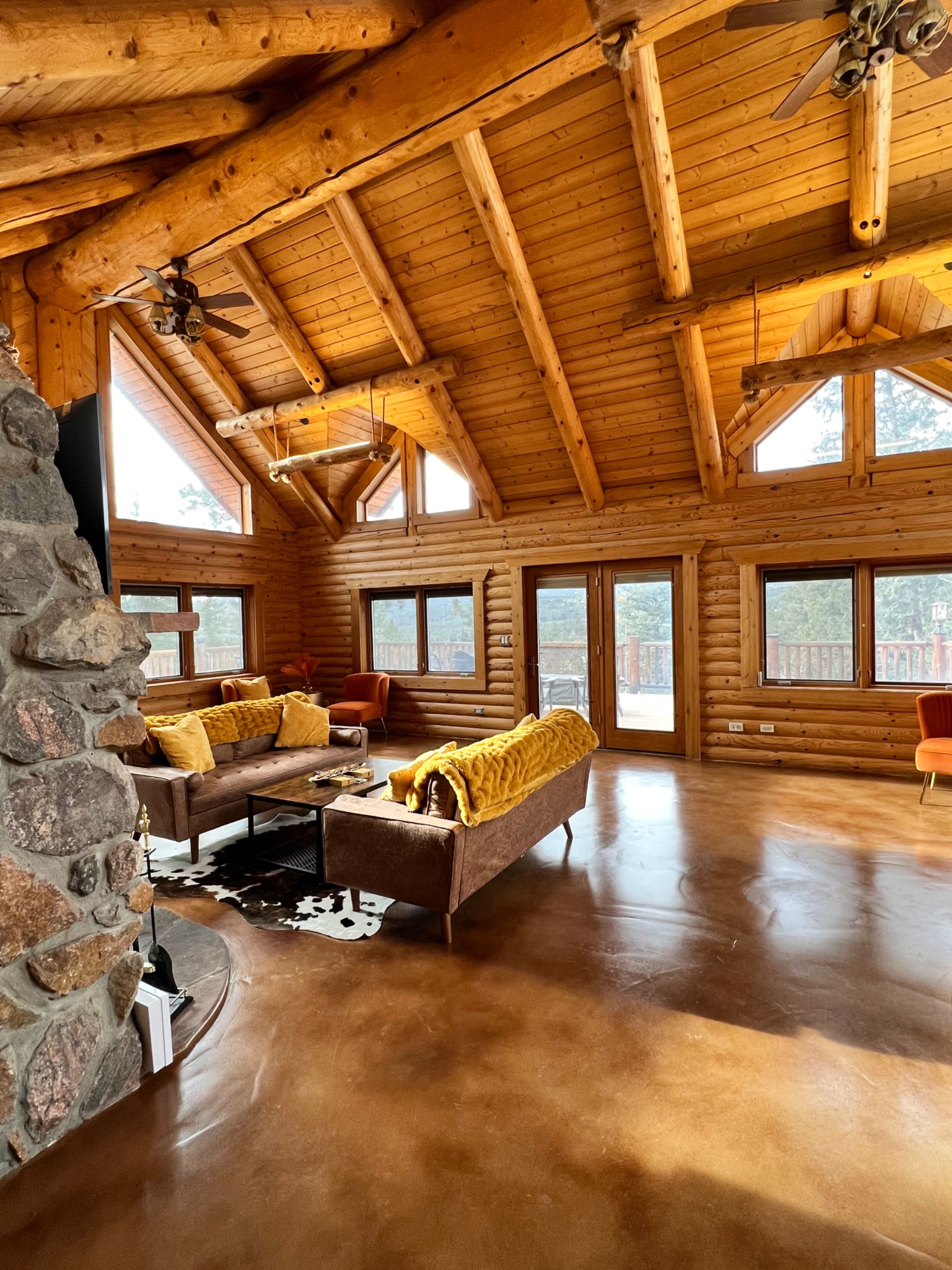 The interior of a spacious log cabin features a high, wooden ceiling with large windows, a polished concrete floor, and cozy seating arrangements.
