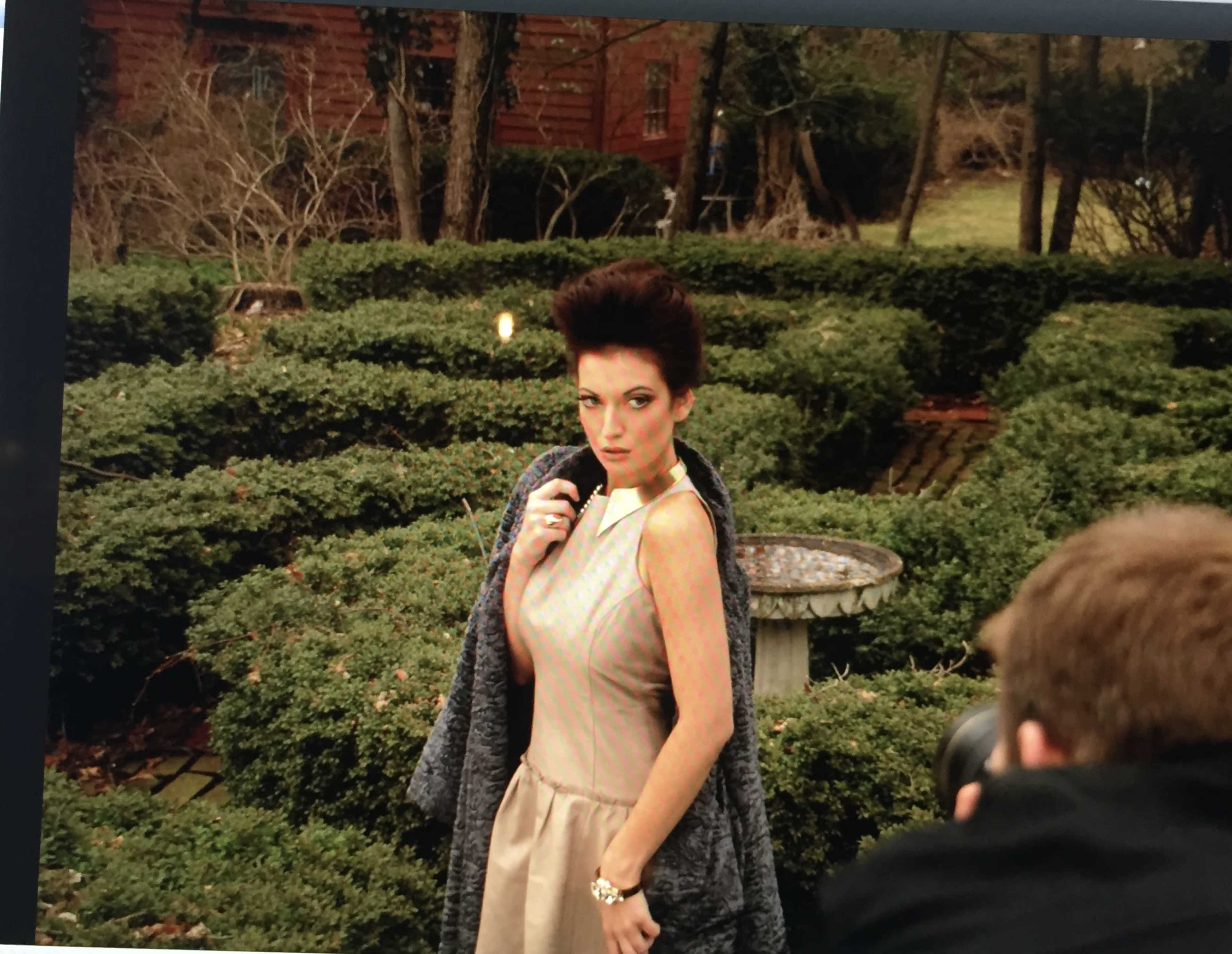 A model poses in a garden with neatly trimmed hedges while a photographer captures her image.