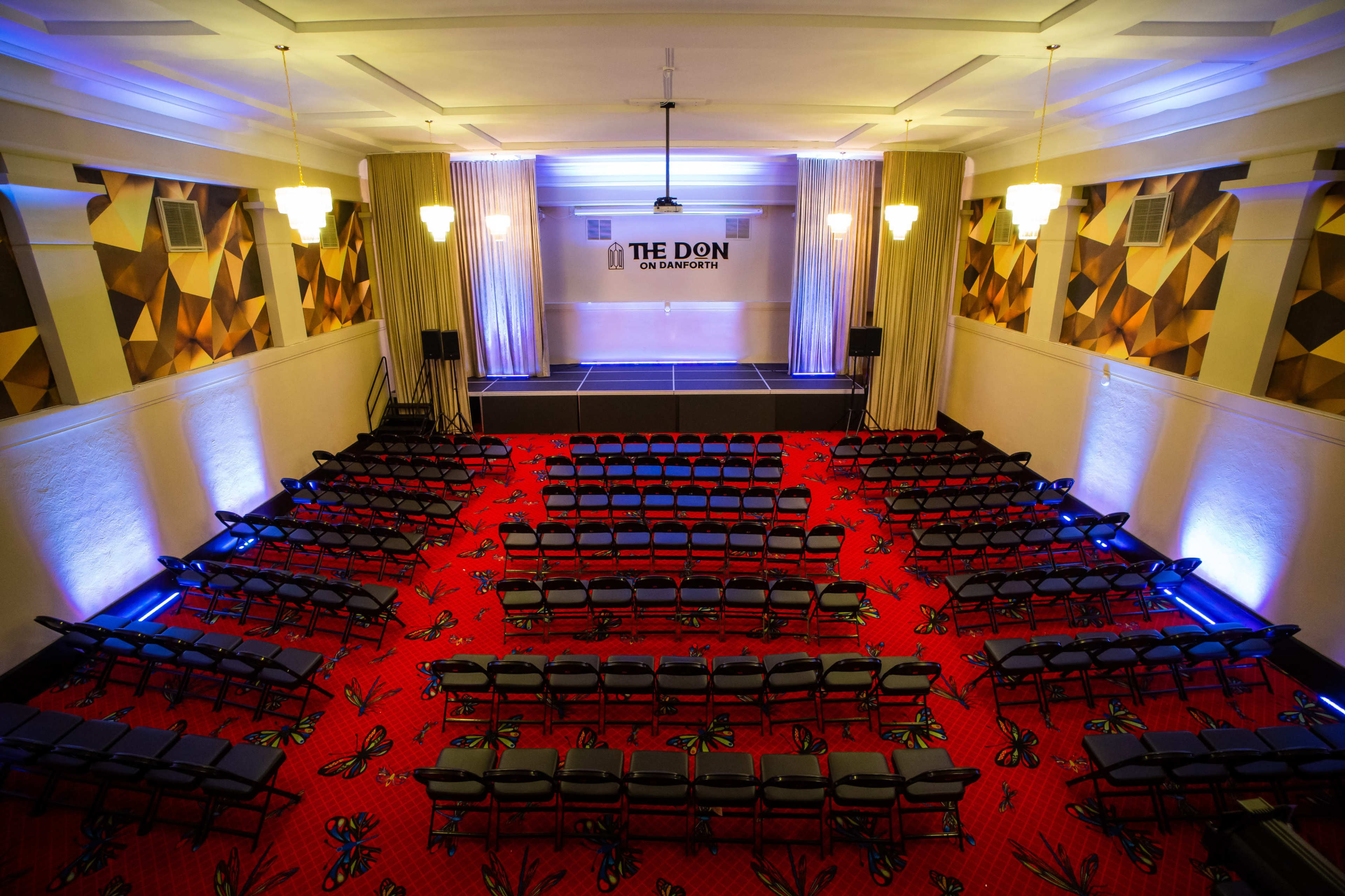 An empty auditorium with rows of black chairs arranged facing a stage, featuring a patterned red carpet and decorative wall treatments.