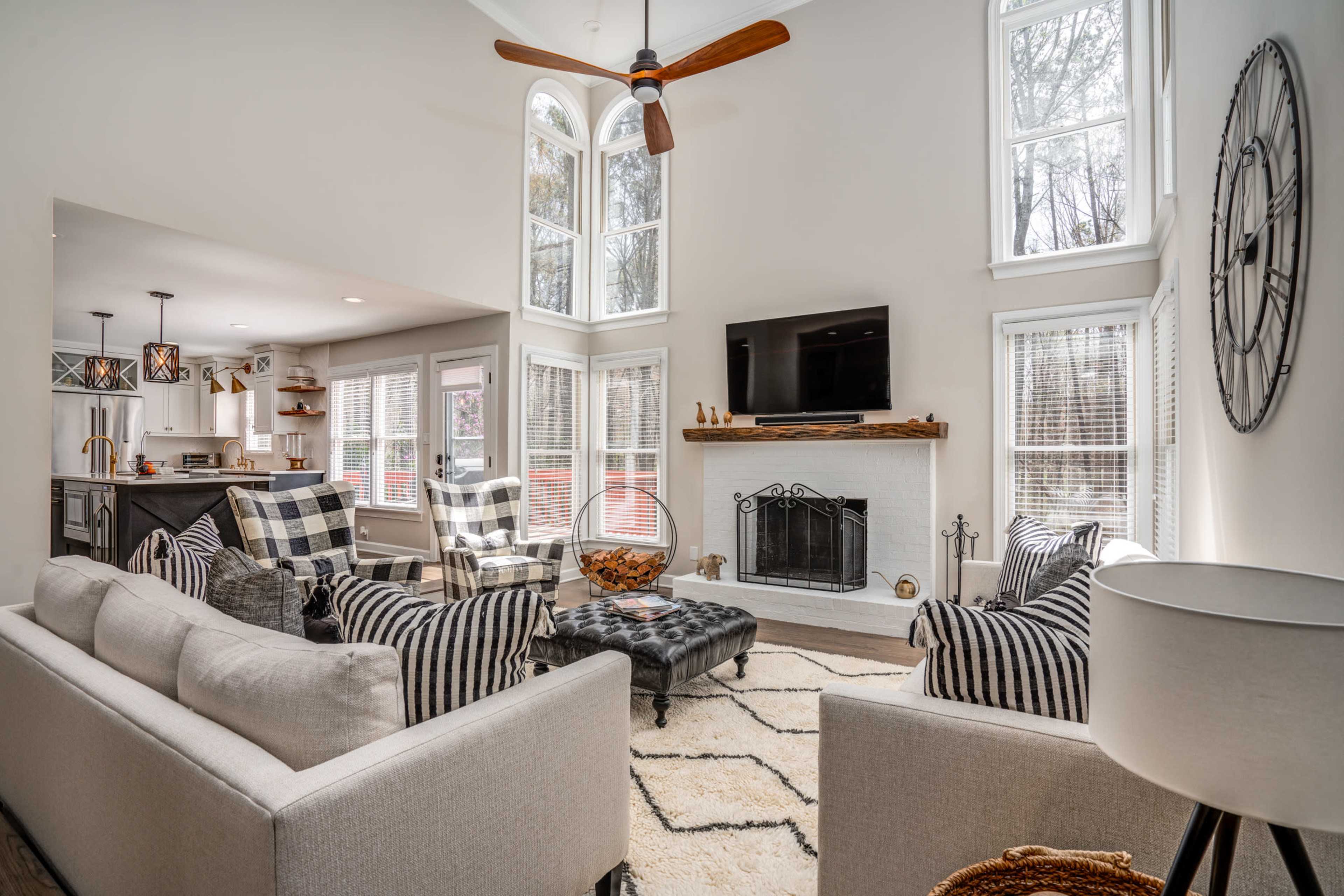 A modern living room features a high ceiling, large windows, a central fireplace, and a mix of patterned furniture arranged around a coffee table.