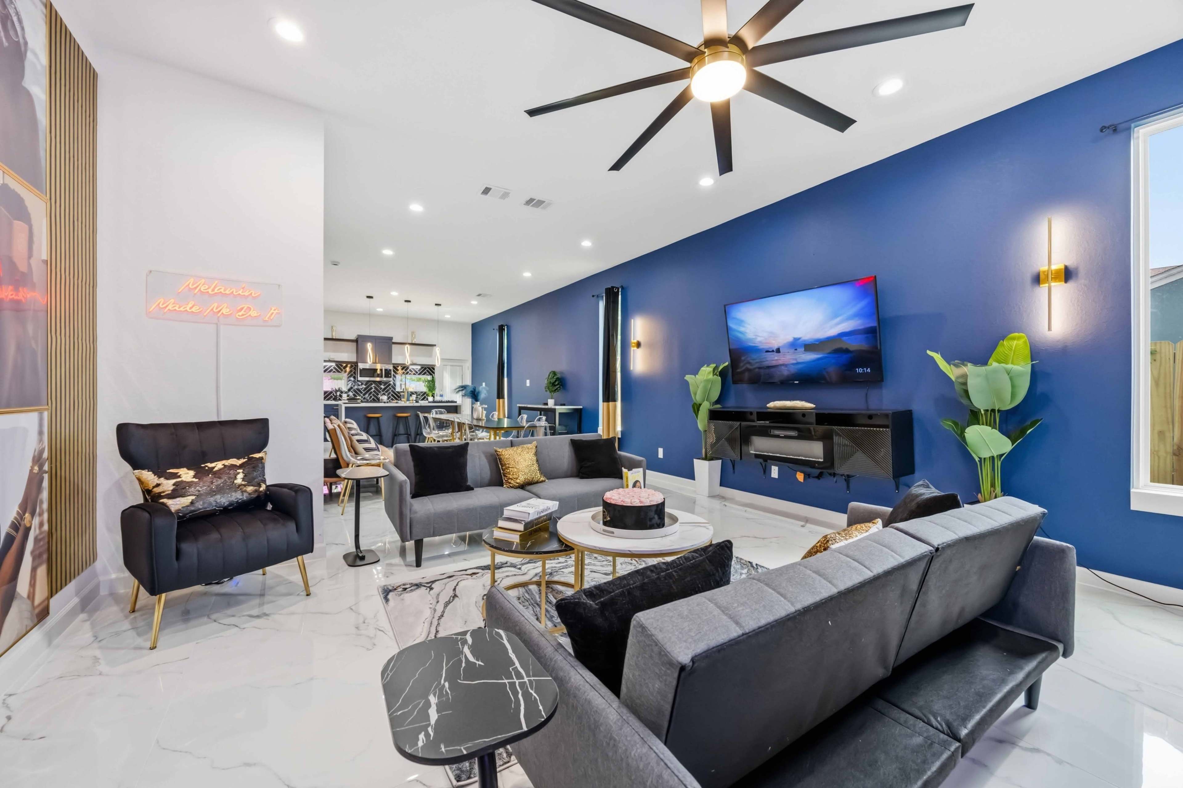 The image shows a modern living room with a blue accent wall, stylish furniture, a large television, and indoor plants.