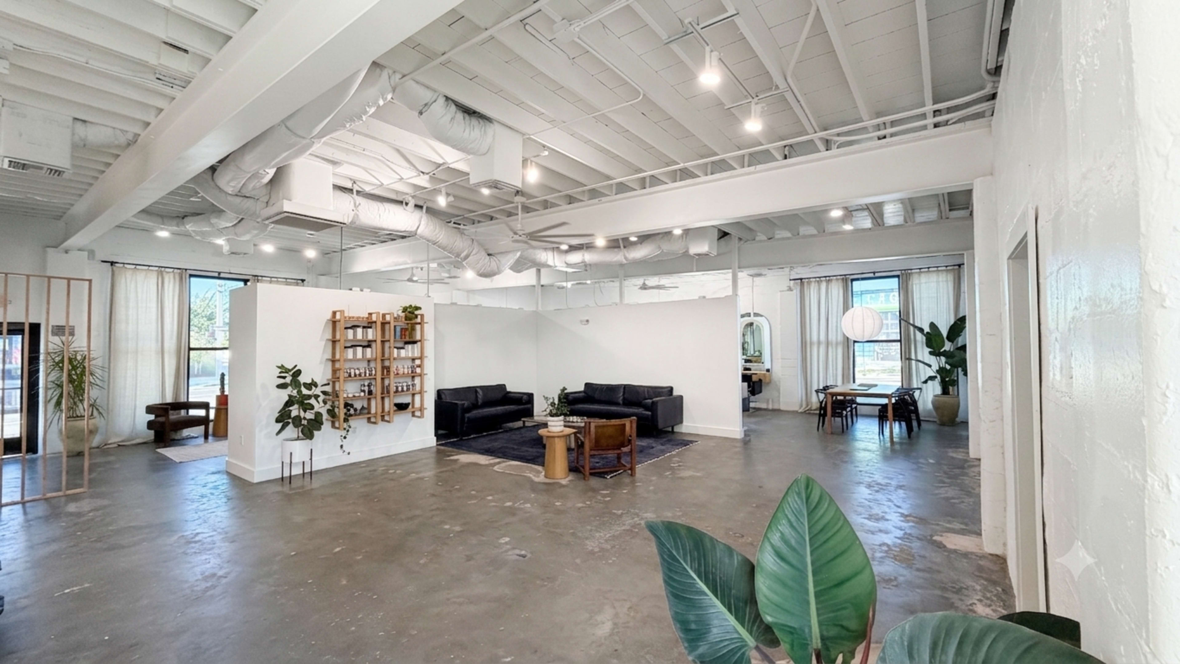 The image shows a spacious, modern interior with various seating areas, a bookshelf, and plants, featuring a combination of natural light and minimalist decor.
