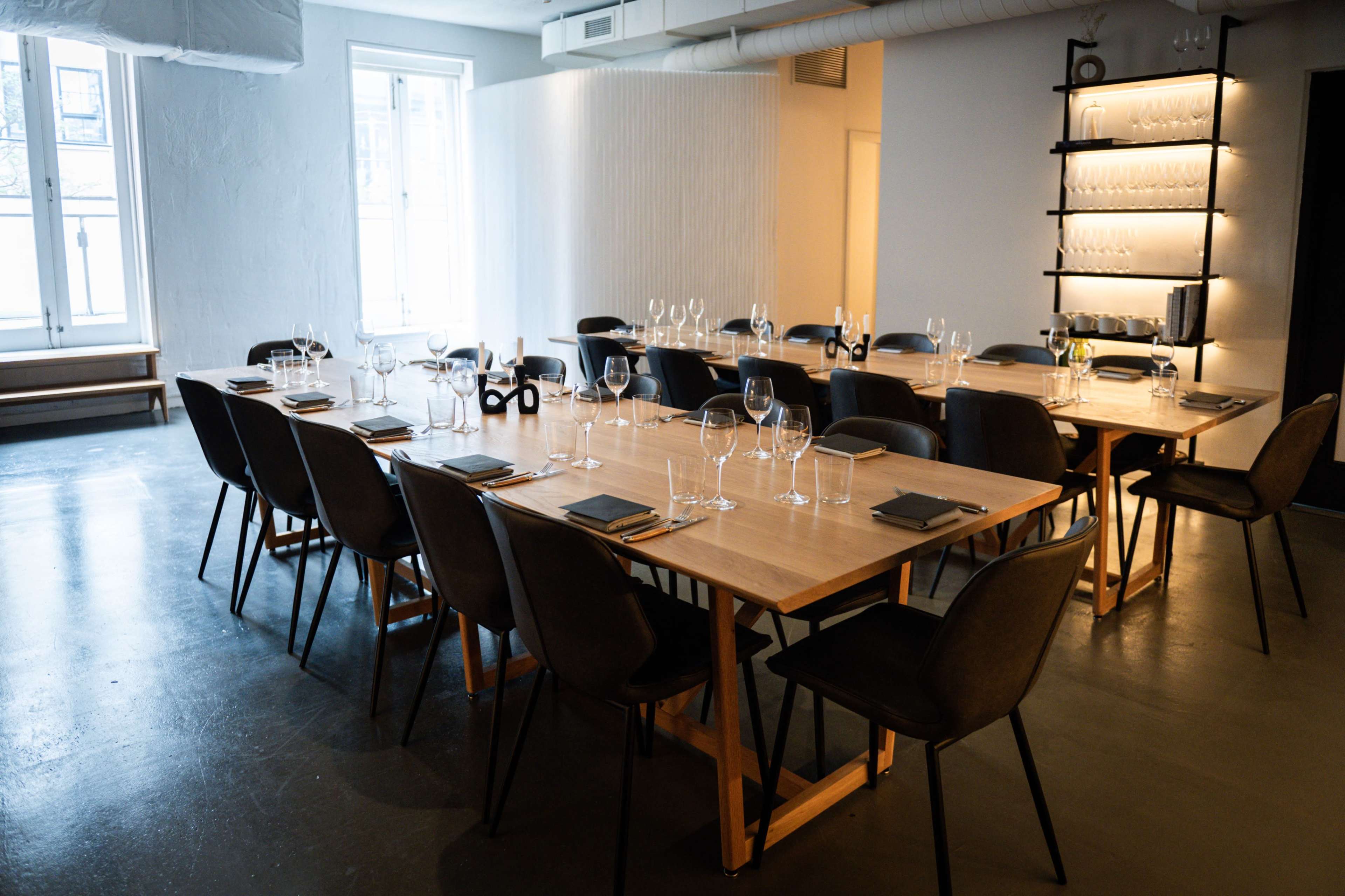 A long wooden table is set for a meal in a spacious dining area with large windows and minimalist decor.