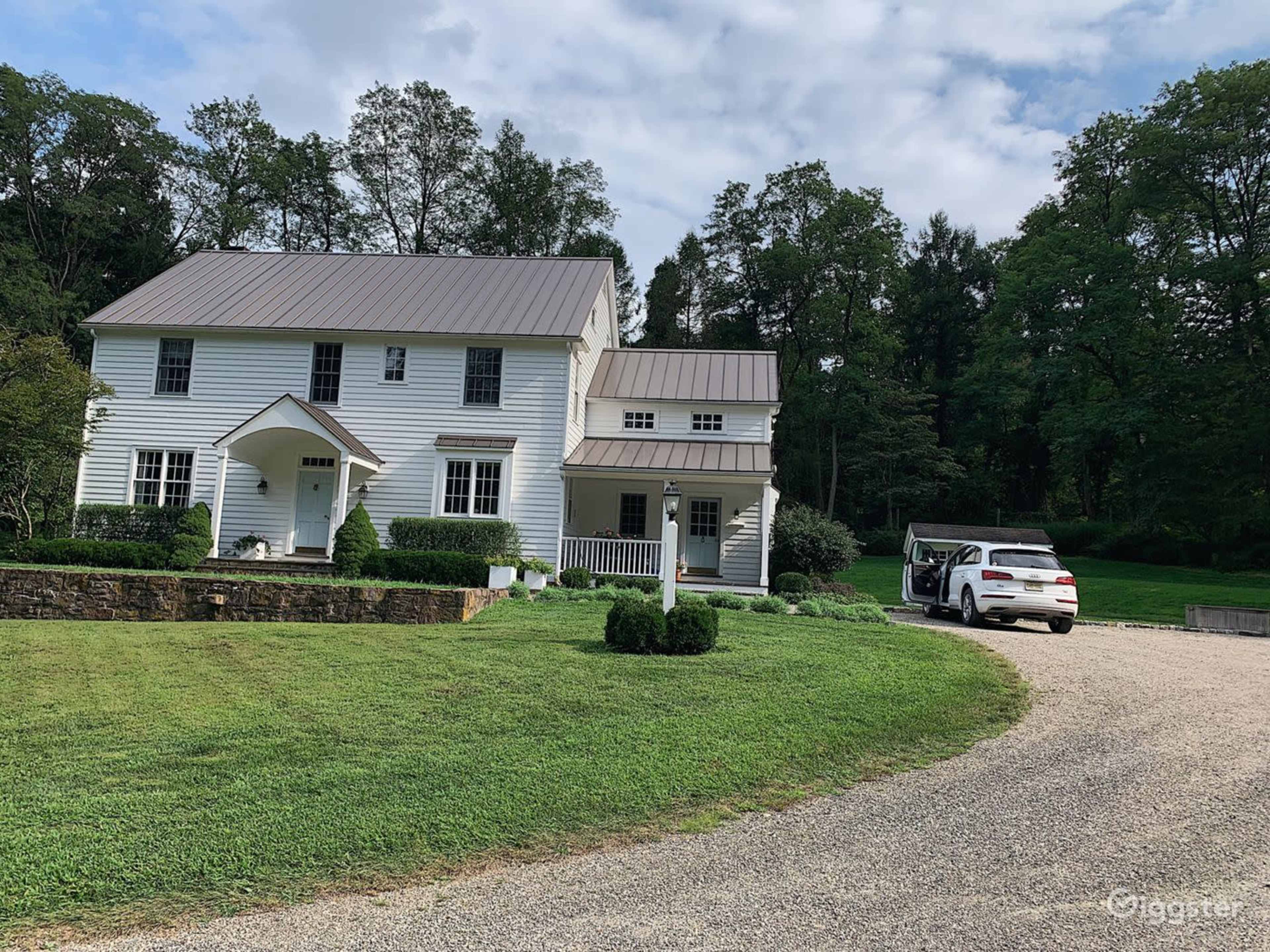A two-story white house with a metal roof is surrounded by grassy areas and trees, with a gravel driveway leading to a parked car.