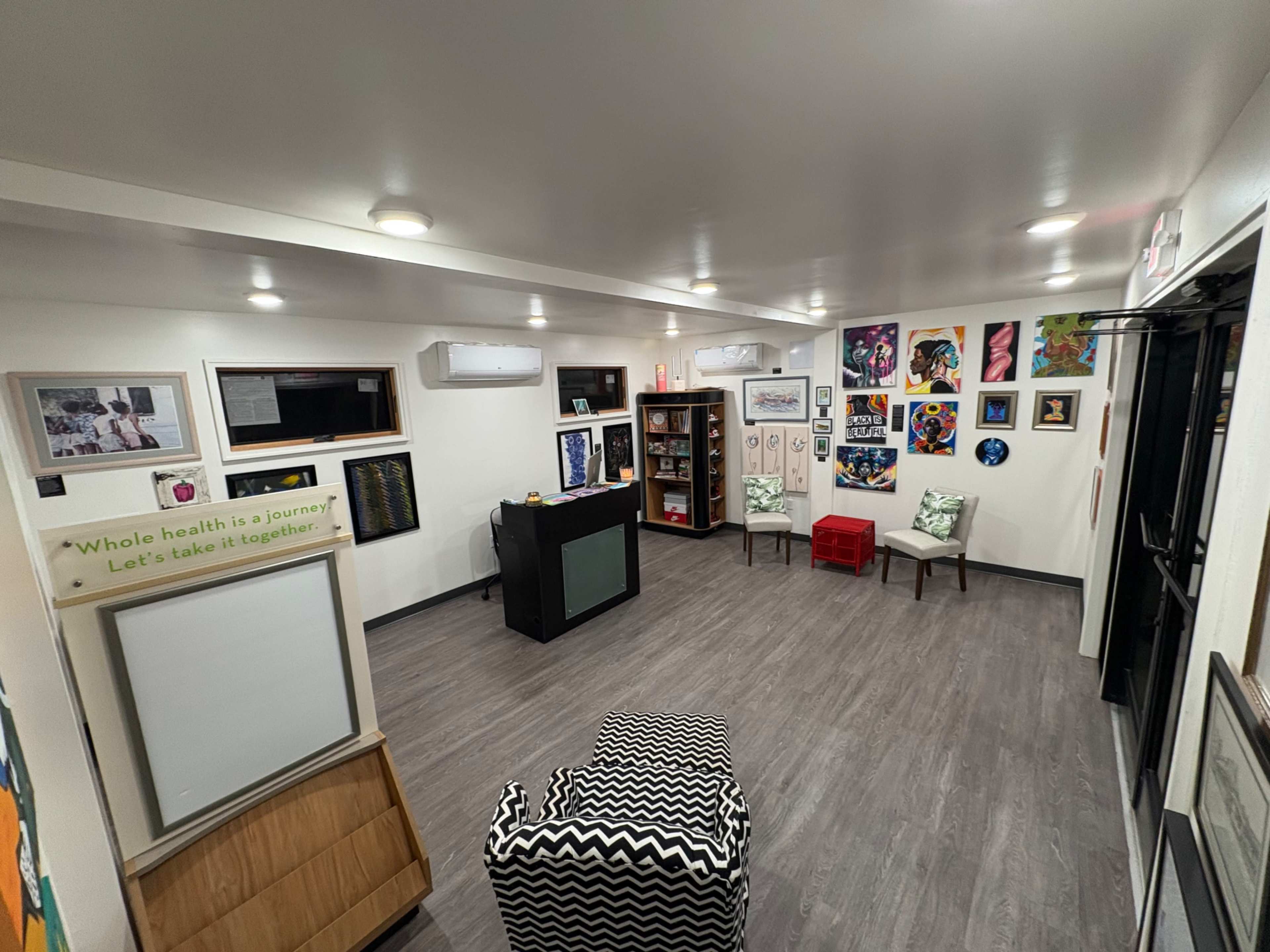 The image shows a modern indoor space with light-colored walls, wooden flooring, and various pieces of artwork displayed, along with seating areas and a reception desk.