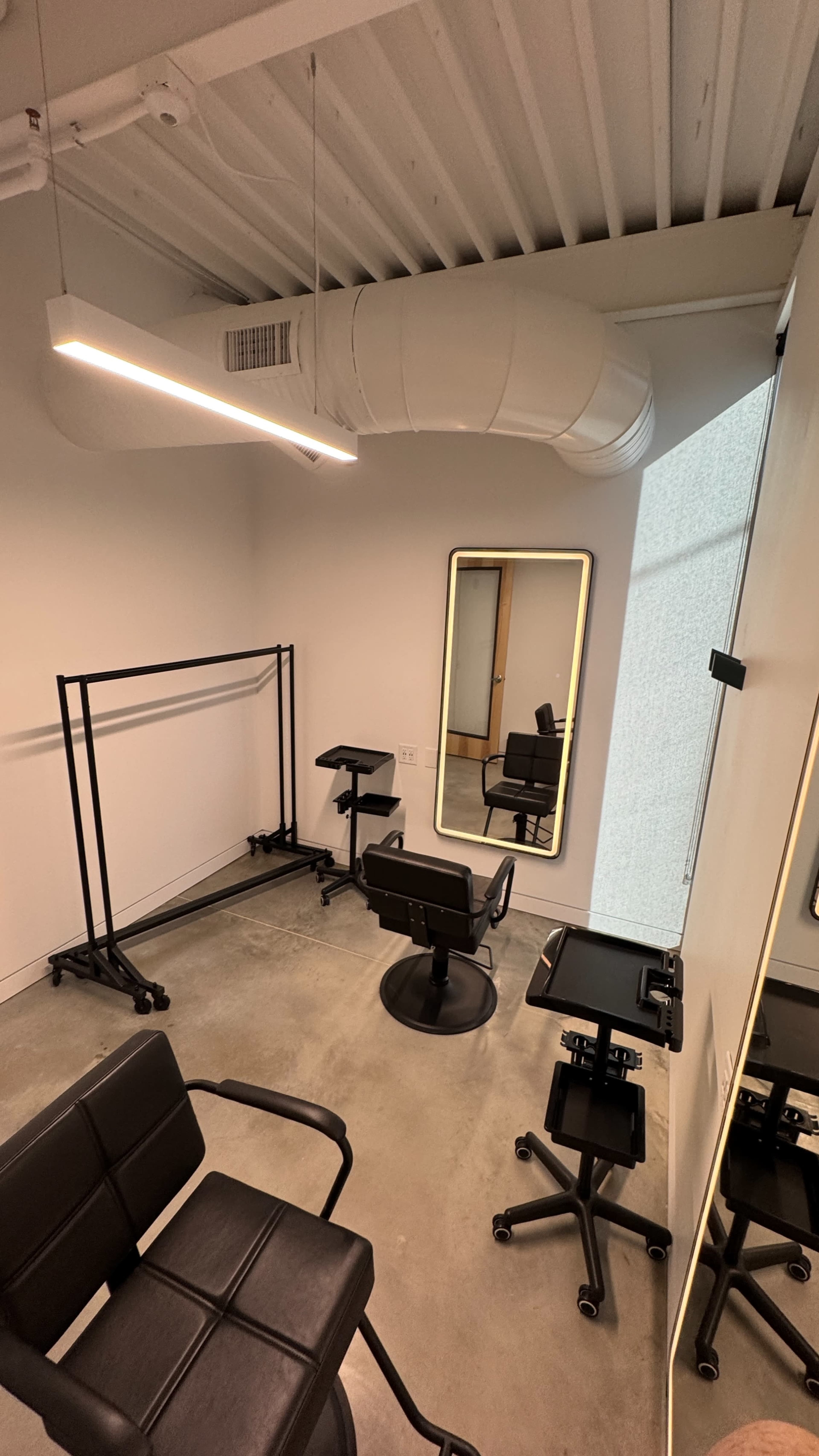 The image shows a minimalist salon interior with two black chairs, a large mirror, rolling carts, and clothing racks against a white wall.
