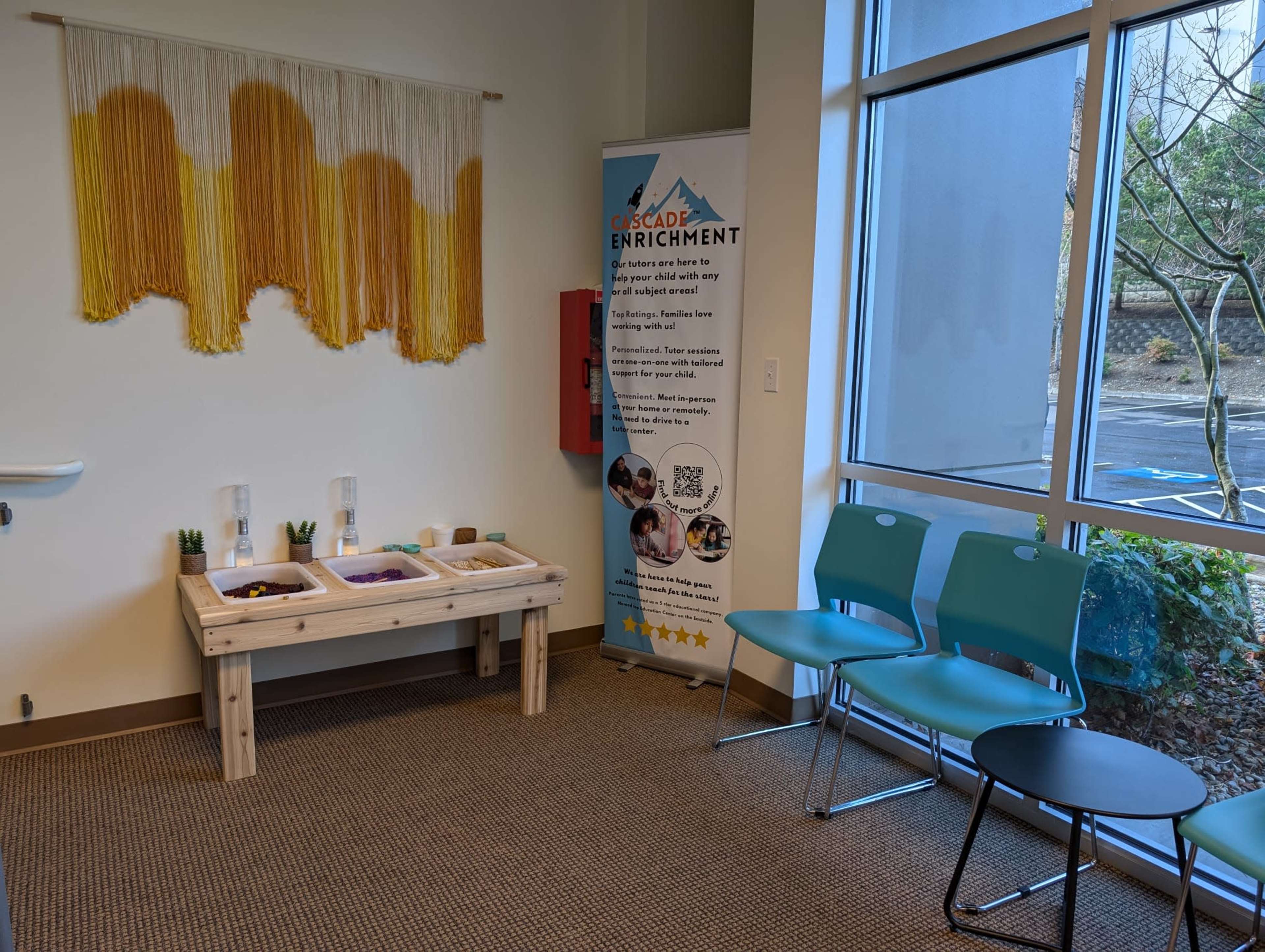 The image shows a waiting area with a wooden table containing bins, a decorative wall hanging, a banner for "Cascade Enrichment," and a few blue chairs arranged nearby.