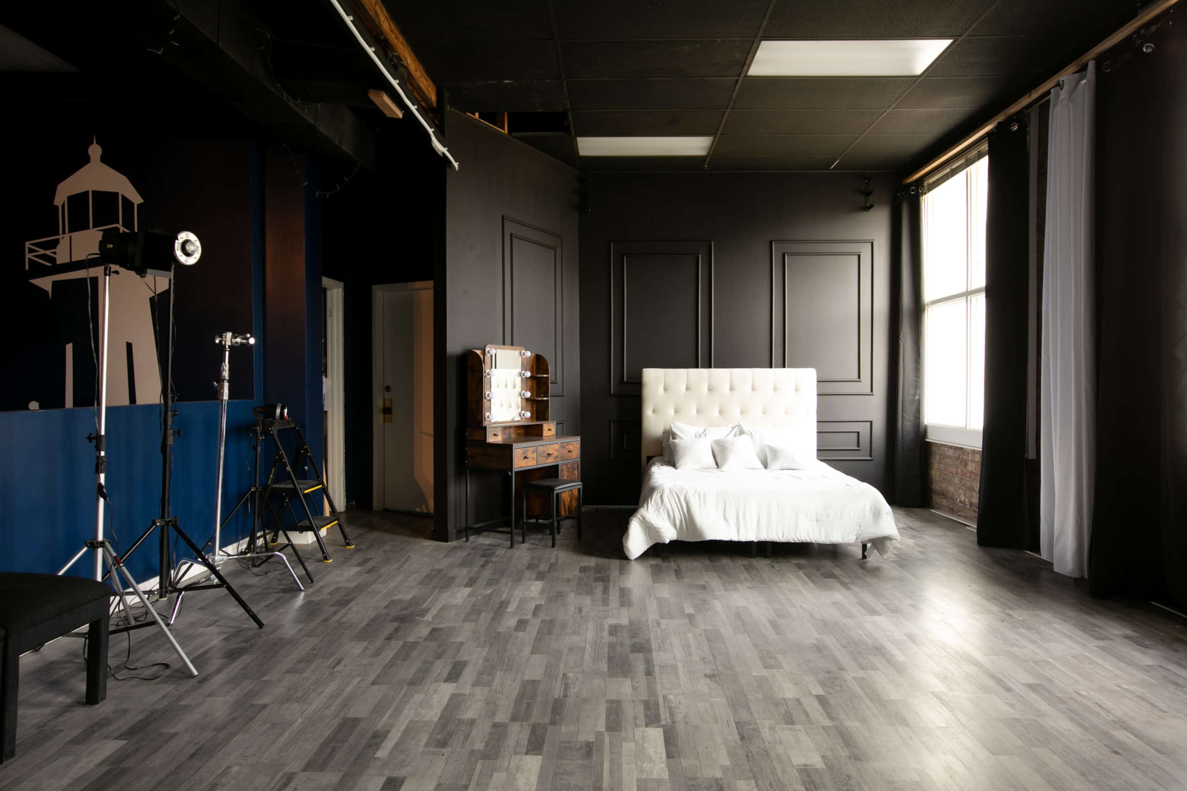 A modern, minimalist bedroom with a white bed surrounded by dark walls and large windows, along with photography equipment set up in the corner.
