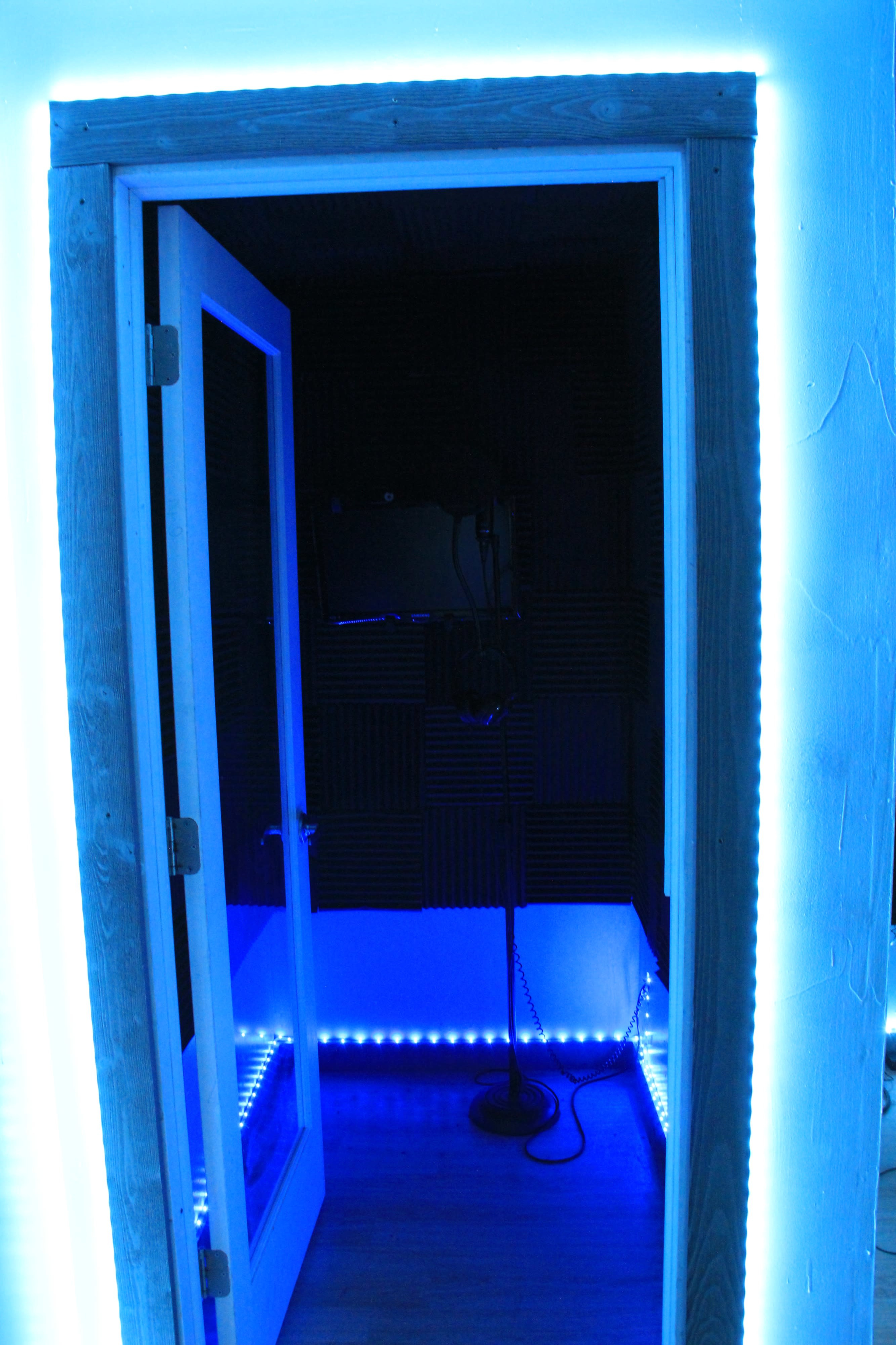 An open door leading into a small, illuminated recording booth with soundproof walls and a microphone stand inside.