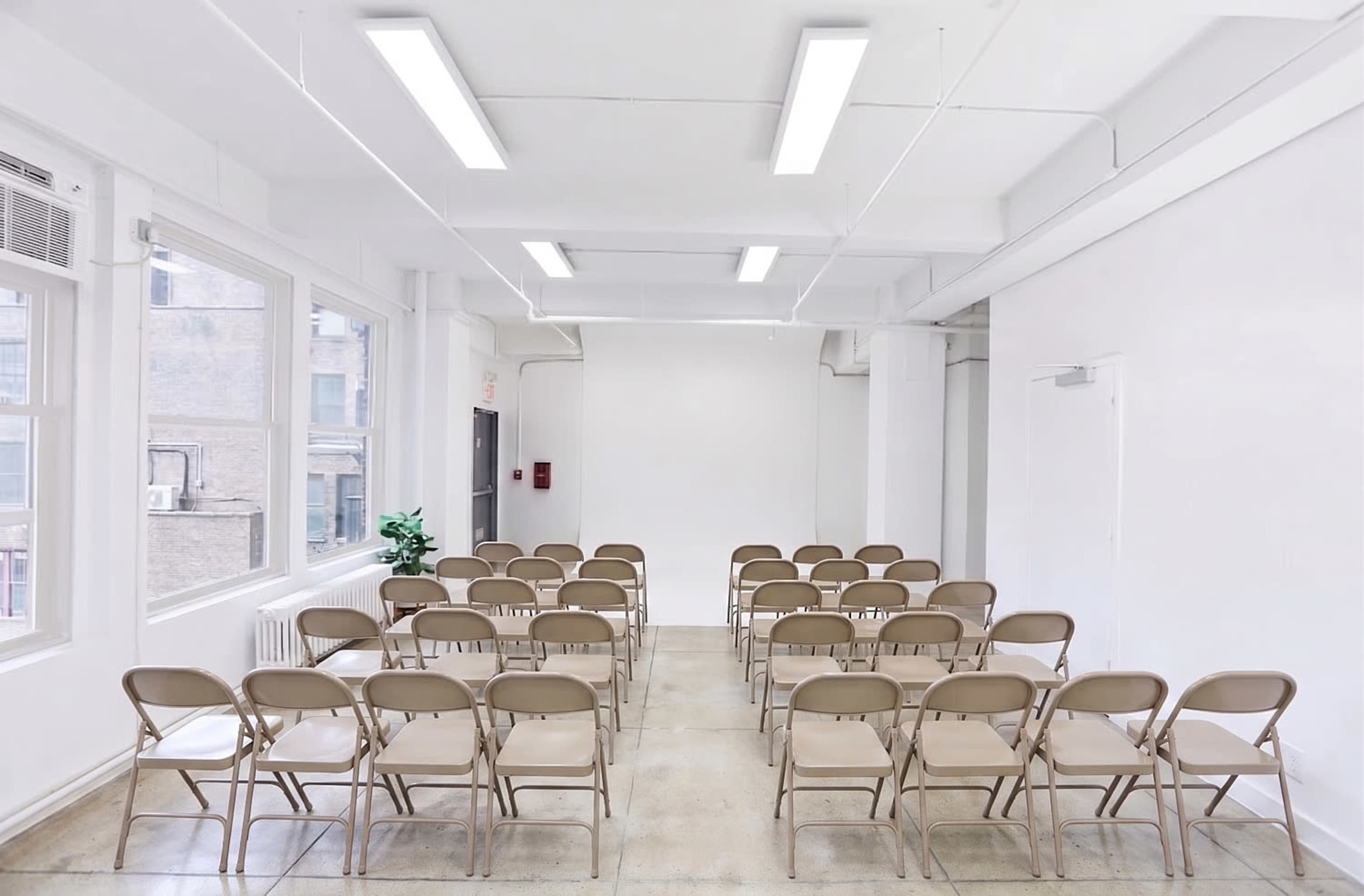 A bright, empty room features rows of beige folding chairs facing a blank wall.
