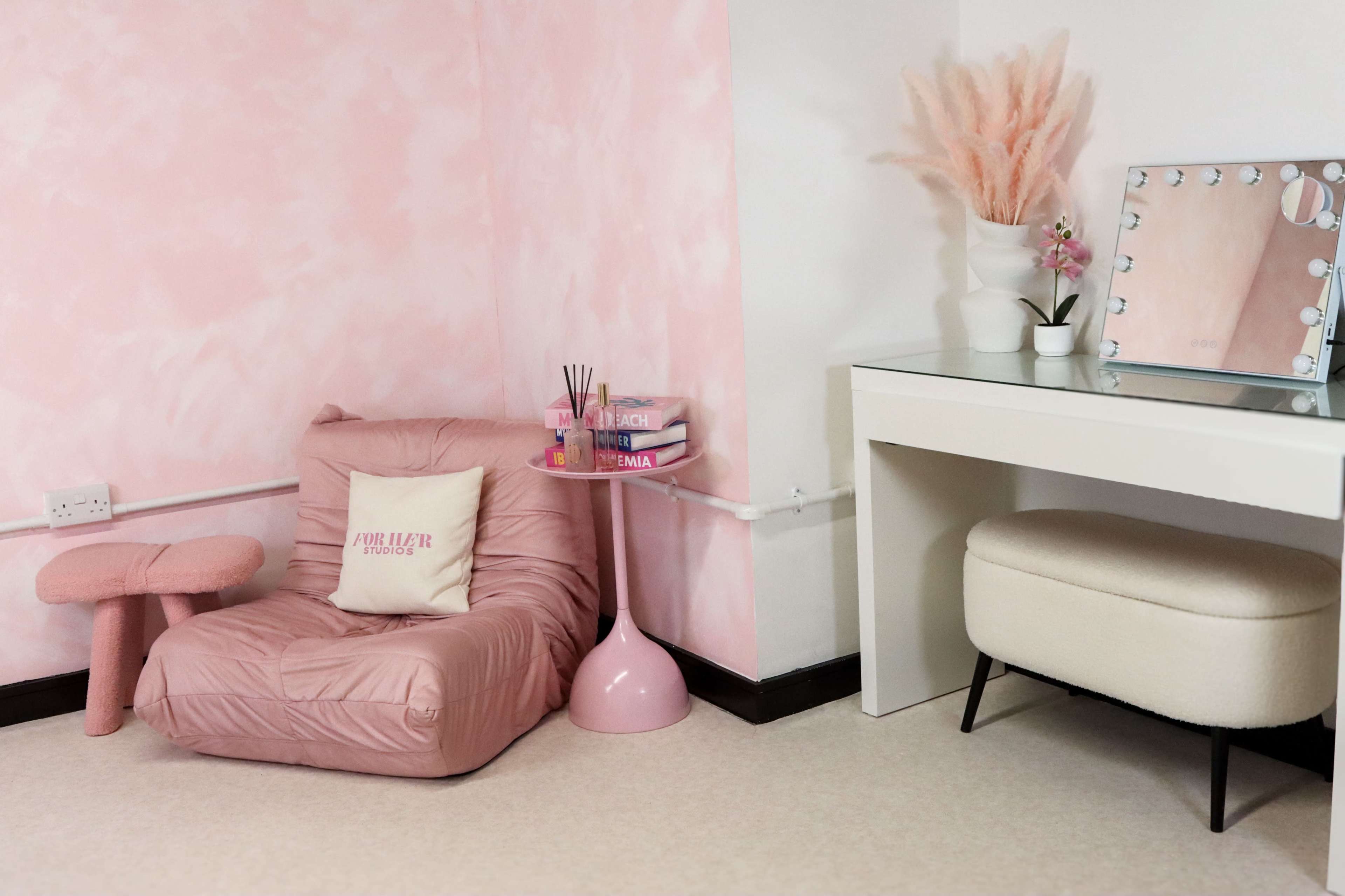 The image features a pink-themed room with a soft chair, a small pink table, and a vanity mirror on a white desk.