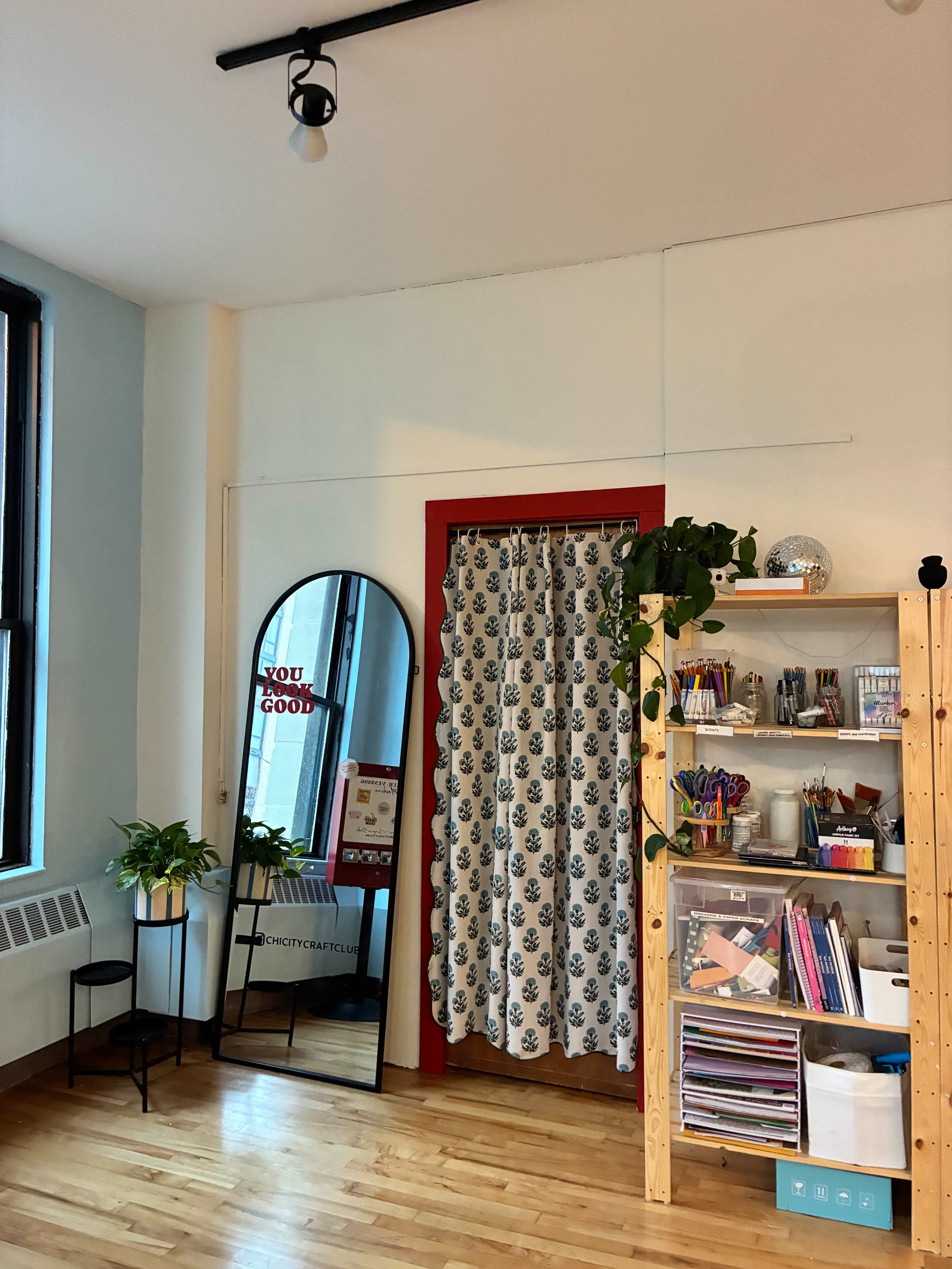 The image shows a brightly lit room featuring a mirror, a red doorway with a patterned curtain, a shelving unit filled with various art supplies, and two potted plants.