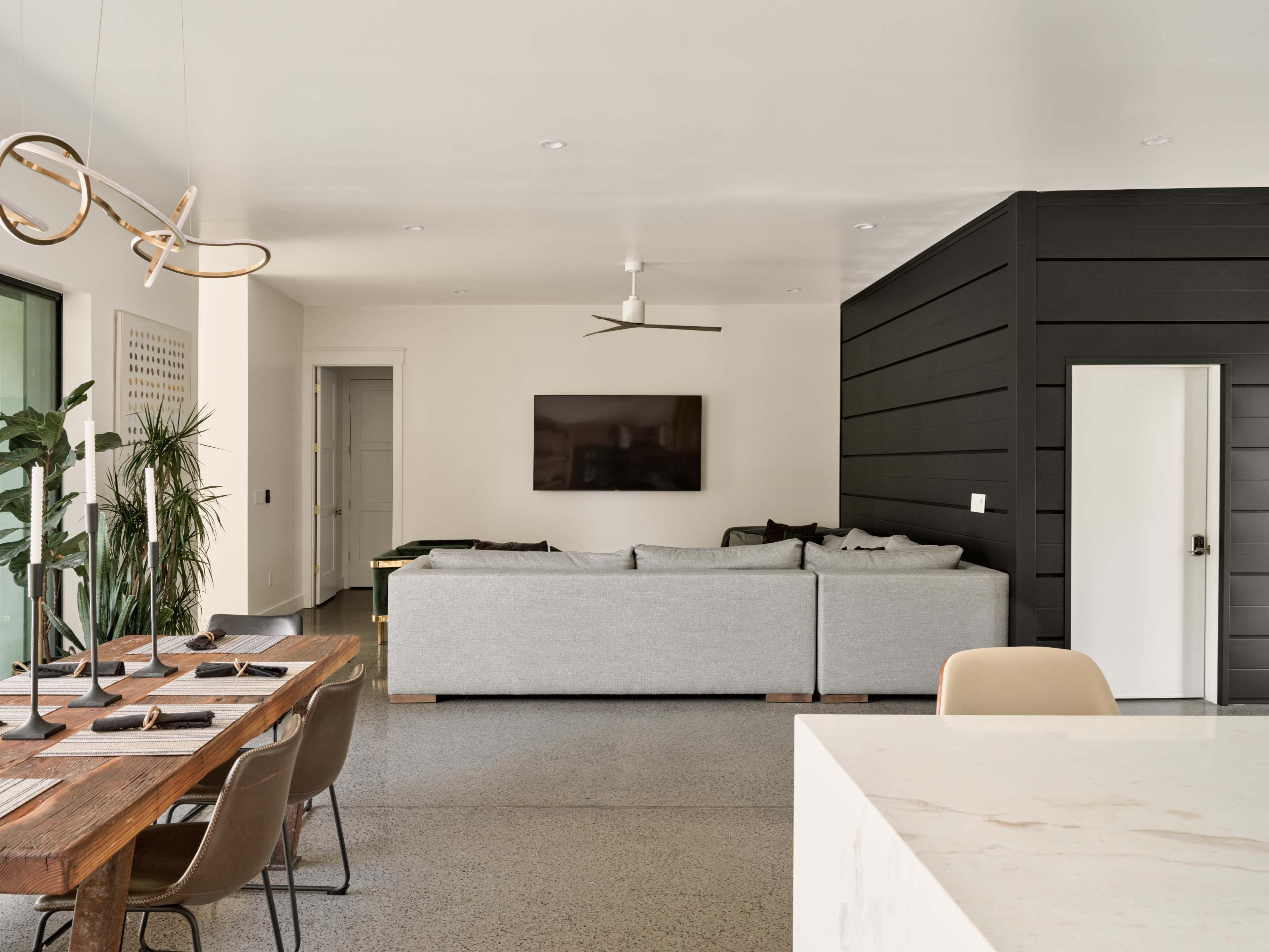The image shows a modern living area featuring a gray sectional sofa, a large flat-screen TV on the wall, a dining table with chairs, and a minimalist kitchen island.