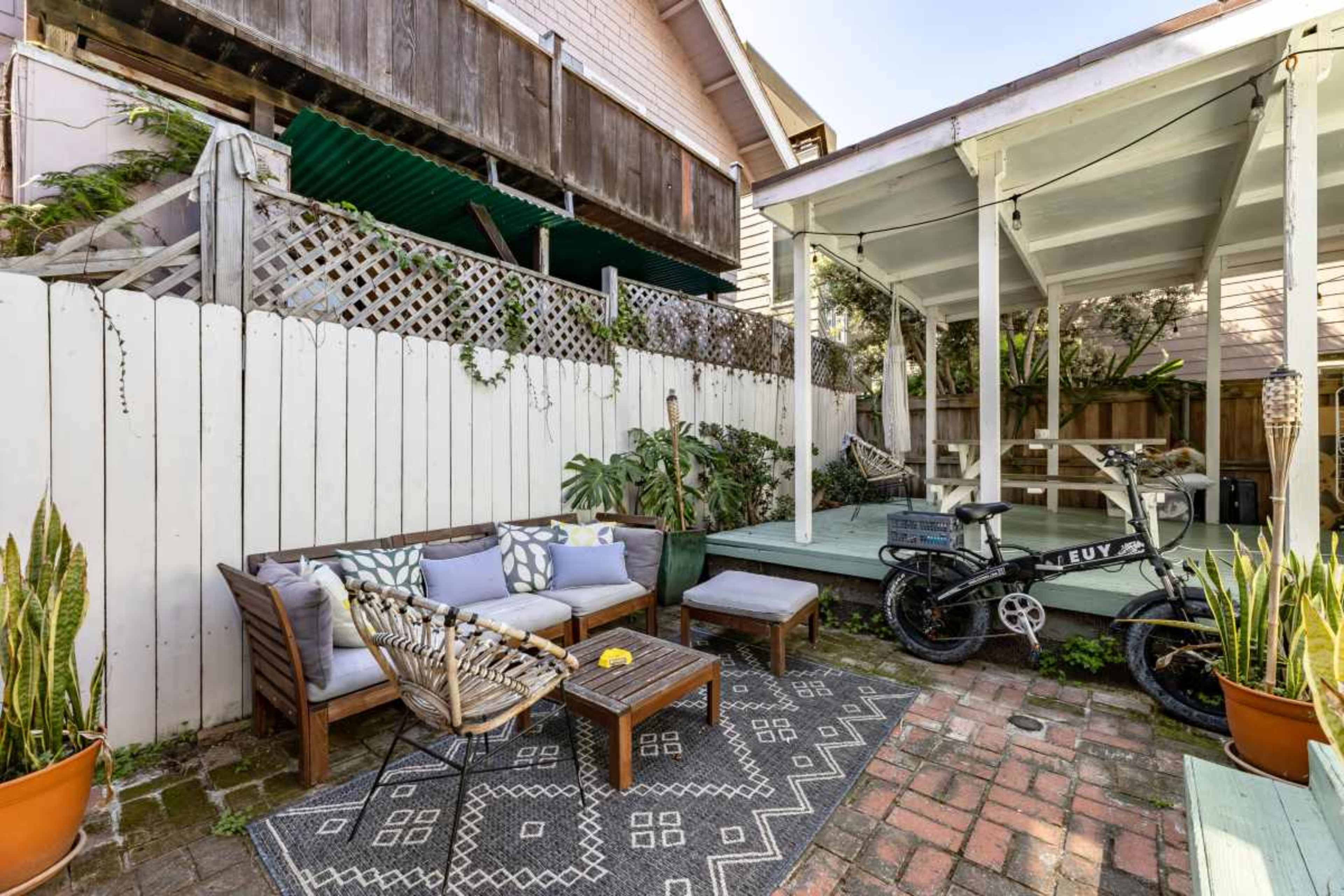 A small backyard featuring a seating area with a wooden couch and a coffee table, surrounded by potted plants and a bicycle parked near a covered patio.