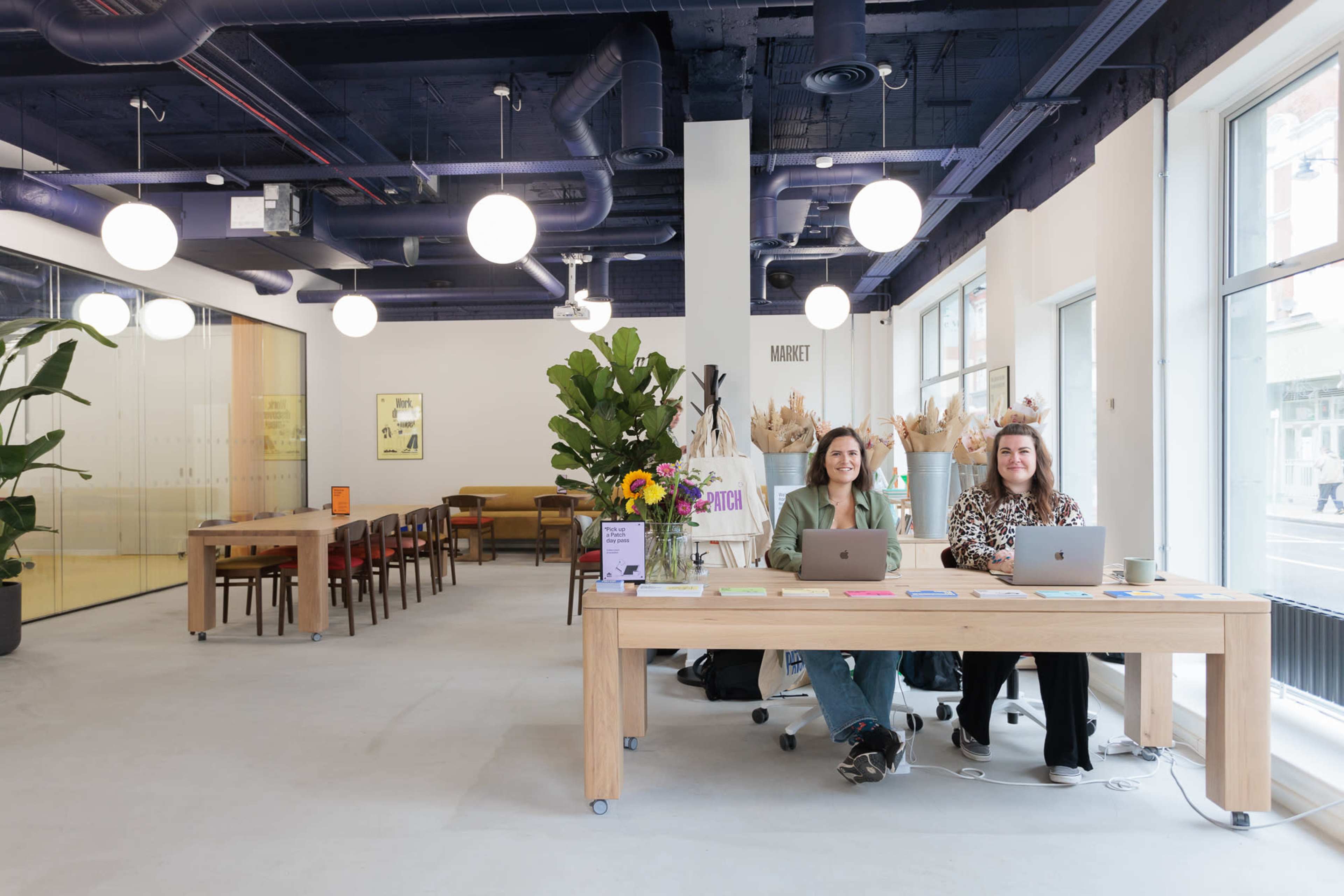 Two women work on laptops at a long wooden table in a bright, modern office space with large windows and minimalist decor.