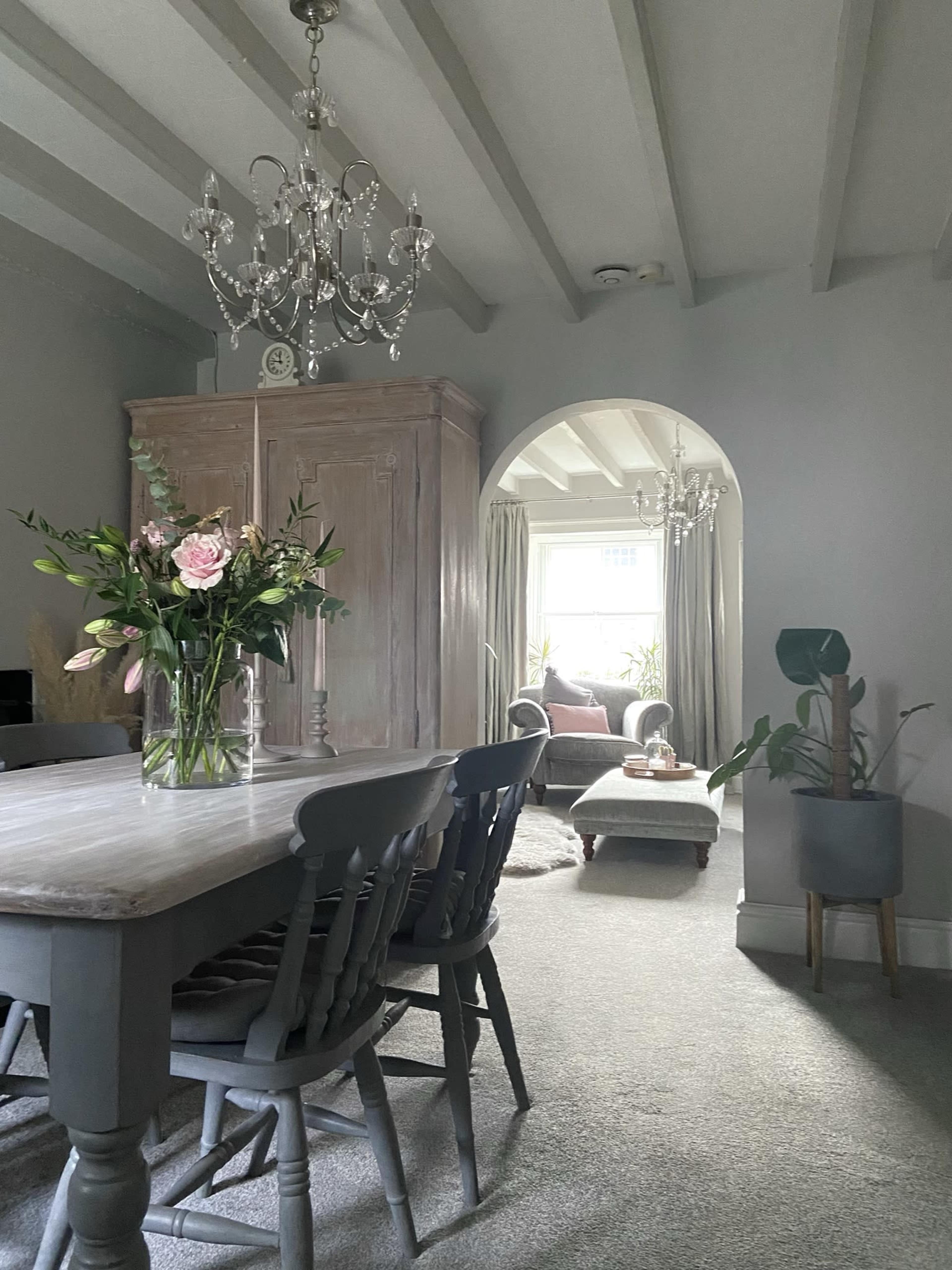 The image shows a dining area with a table and chairs, a flower vase, and an archway leading to a cozy lounge with a sofa and decorative plants.