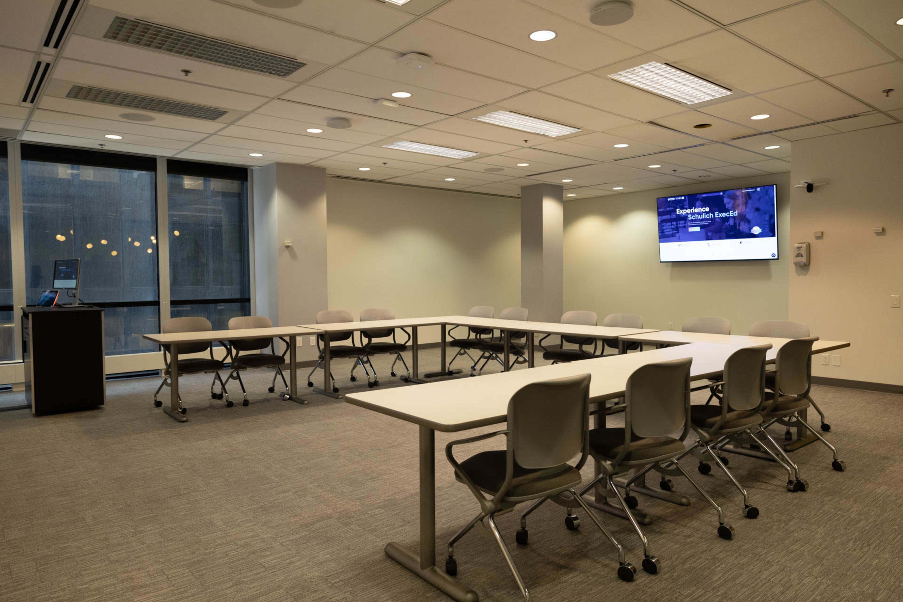 A modern conference room features a large table surrounded by chairs, a wall-mounted screen, and windows providing natural light.