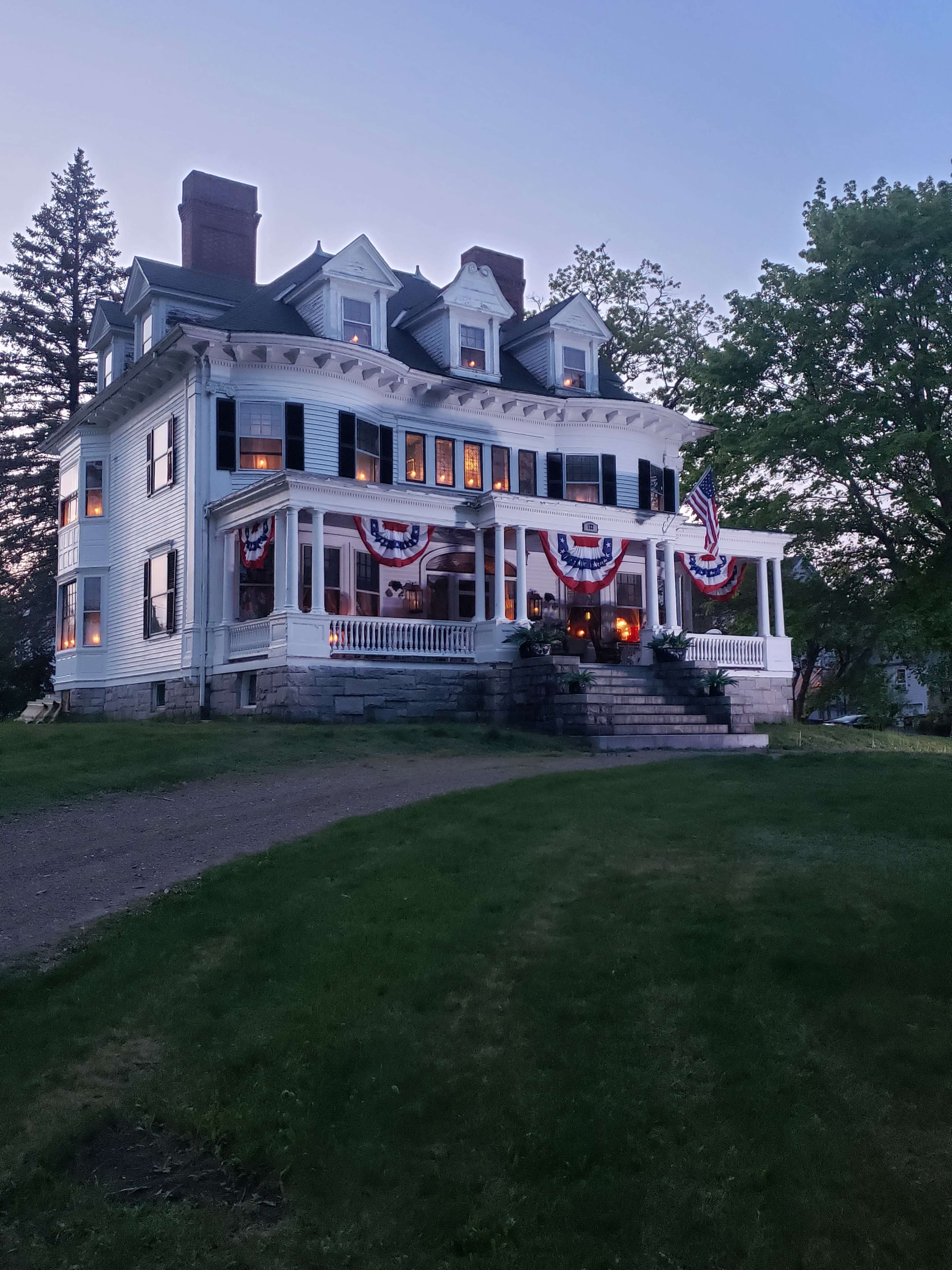 A large, three-story white house with multiple windows and a porch is decorated with red, white, and blue bunting and has lights visible inside.