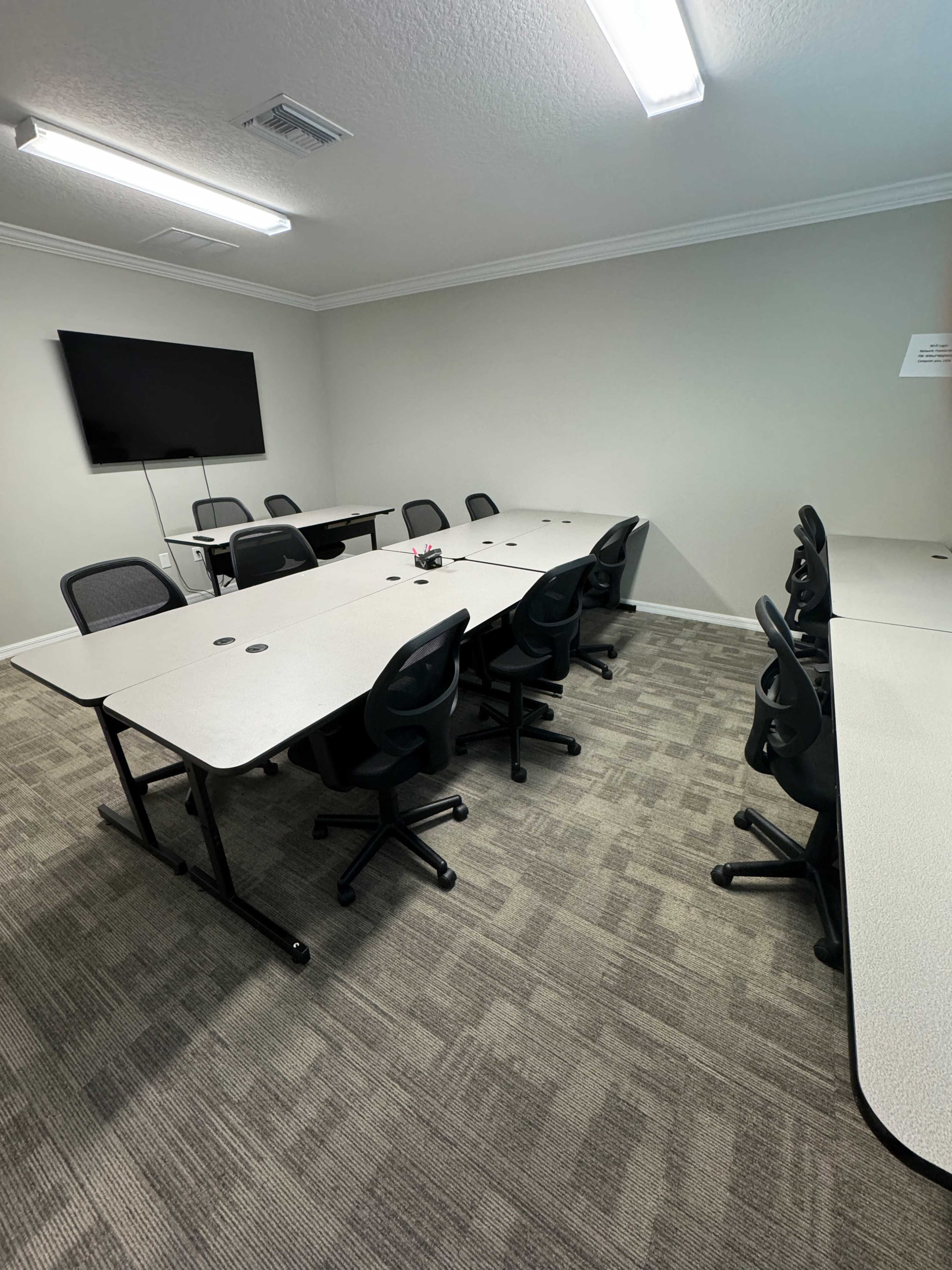 A conference room is shown with several tables arranged in a U-shape, surrounded by black swivel chairs and featuring a wall-mounted TV.
