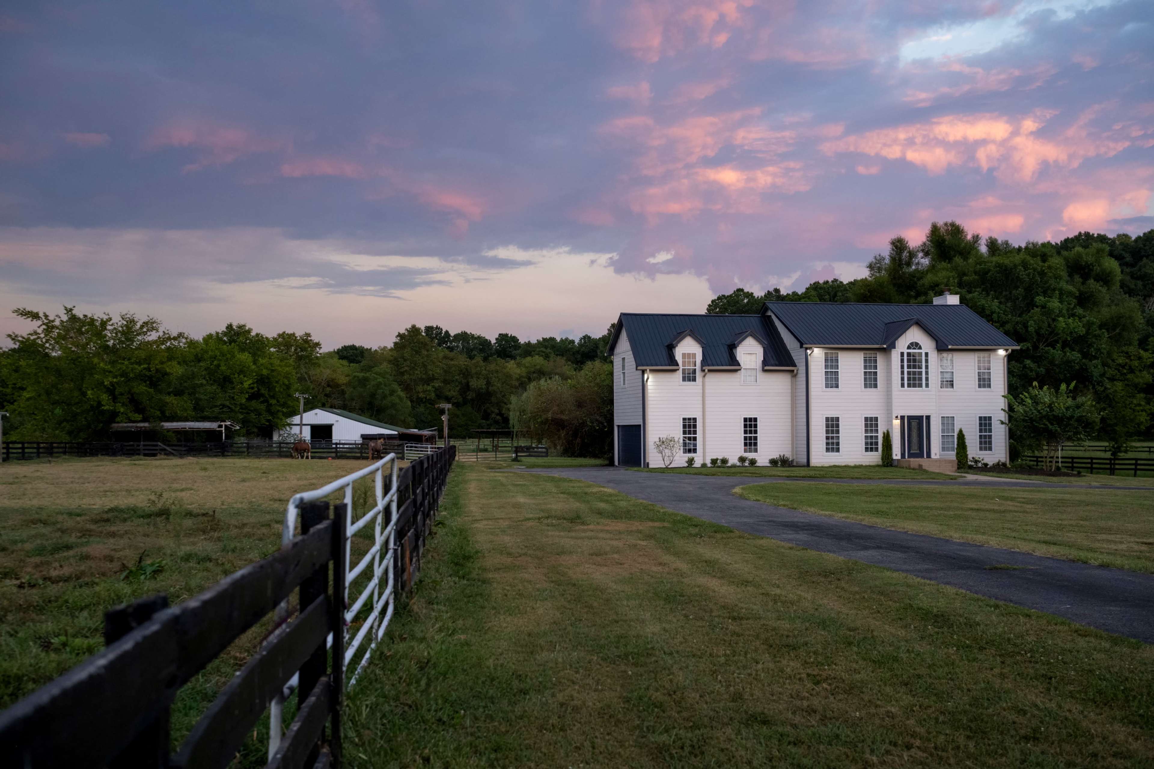 A white house with a dark roof stands next to a gravel driveway, bordered by a wooden fence and surrounded by green fields under a colorful sunset sky.