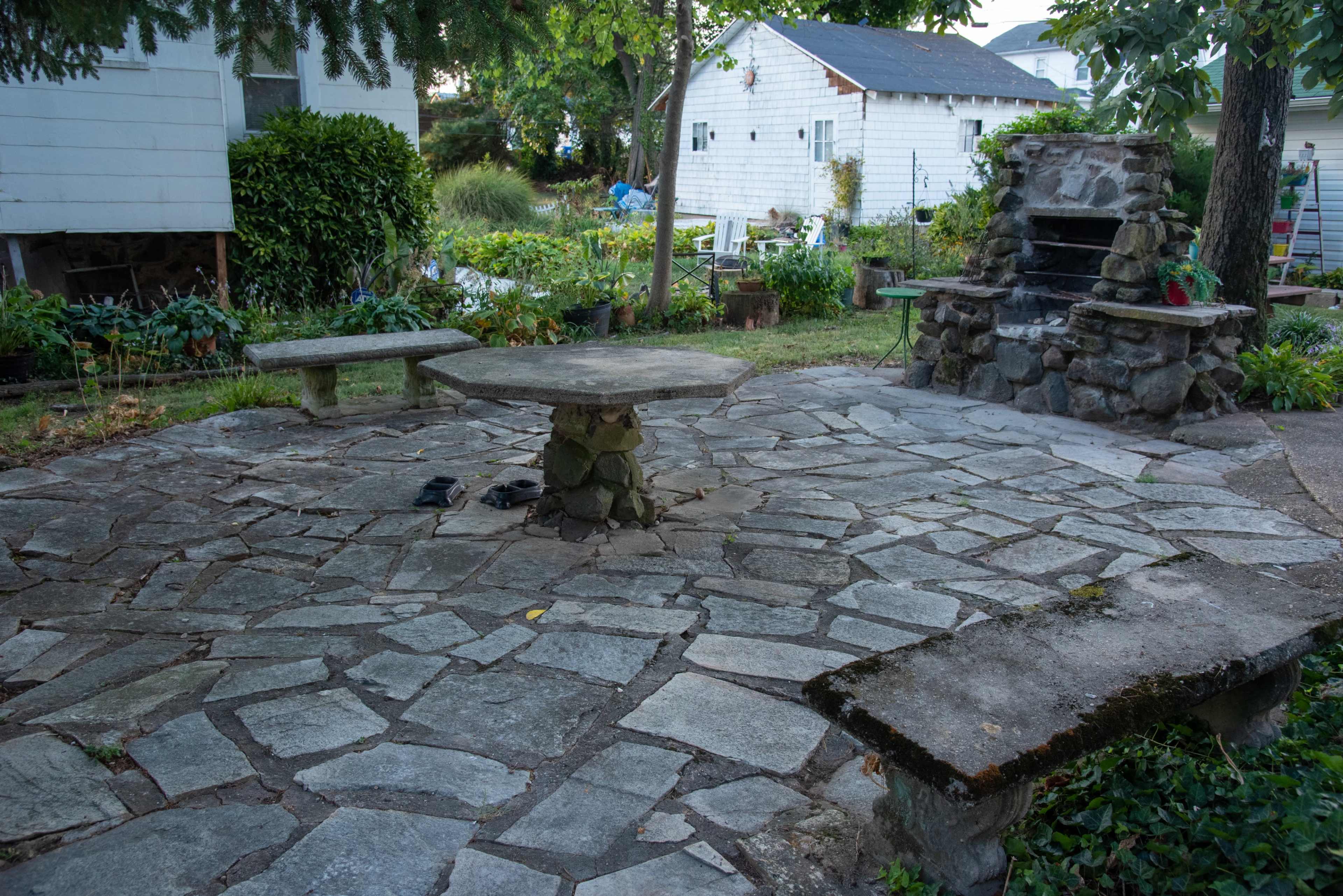 A stony patio with a central table and surrounding benches leads to a stone fireplace, set in a garden with various plants in the background.