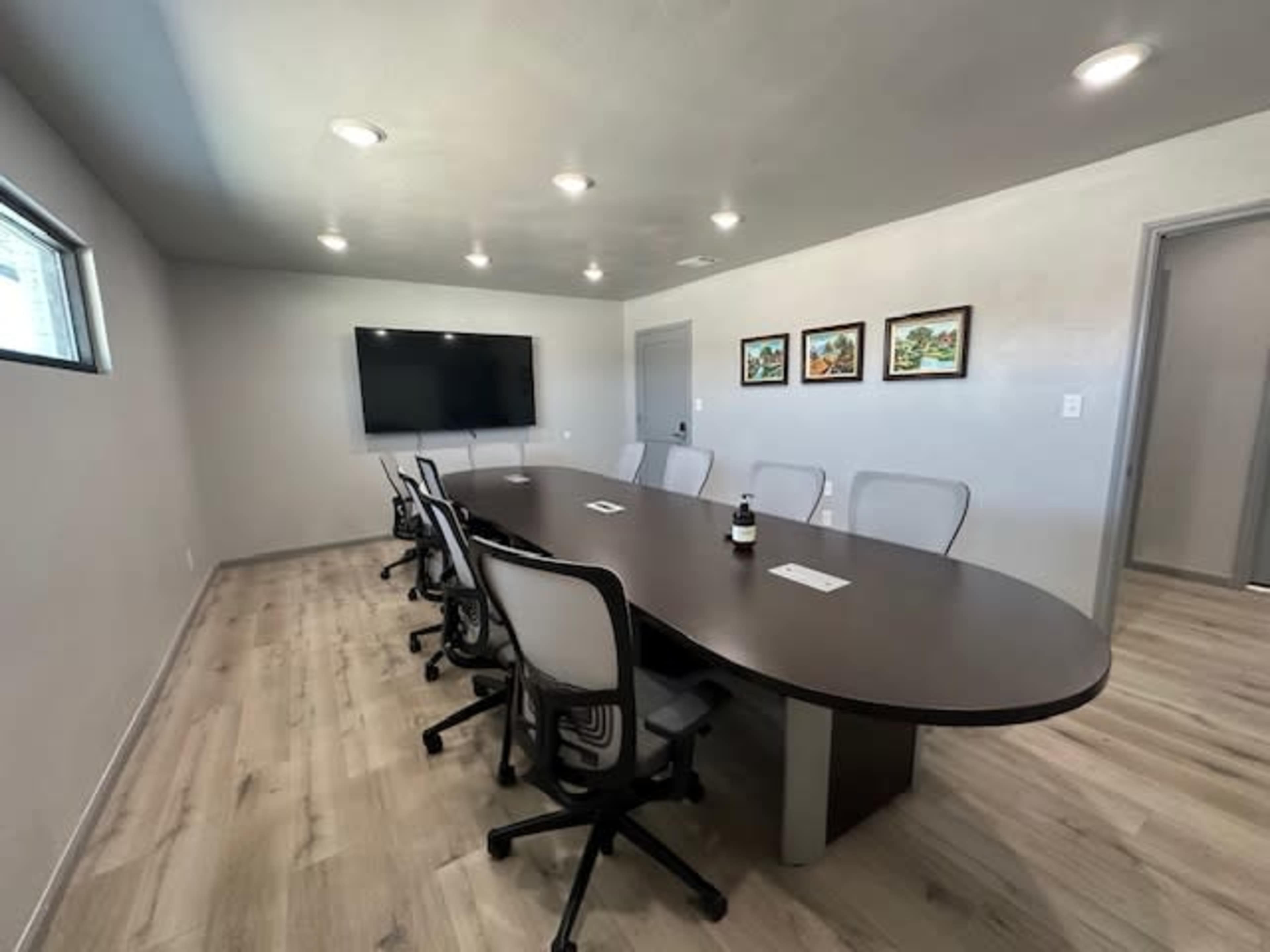 The image shows a modern conference room featuring a large table, chairs, a television, and framed artwork on the walls.