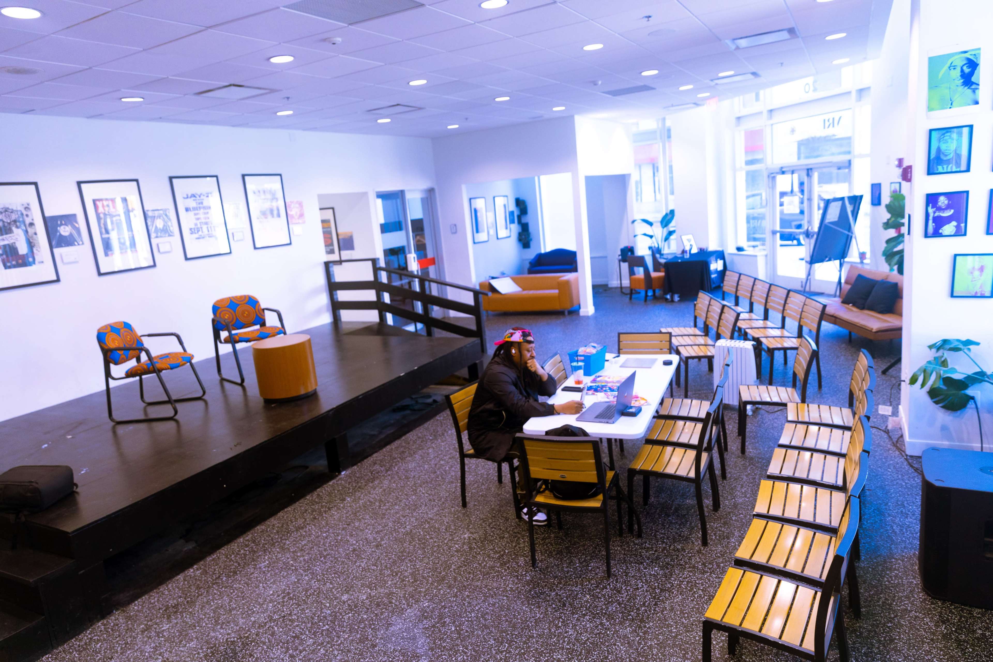 A person sits at a table in a spacious room featuring a performance stage, rows of chairs, and various artworks on the walls.