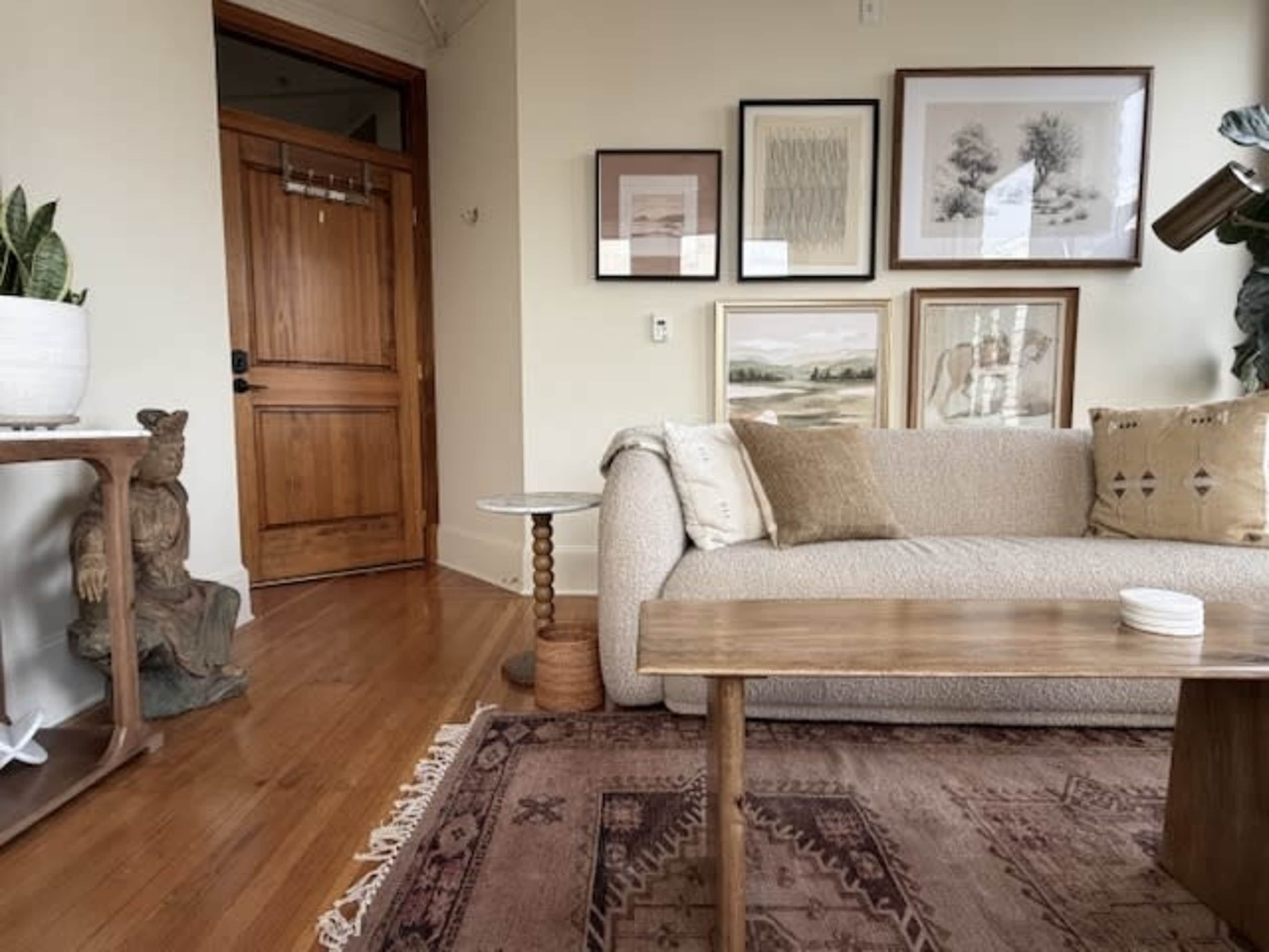 The image shows a cozy living room featuring a light-colored sofa, a wooden coffee table, framed artwork on the walls, and a wooden front door.