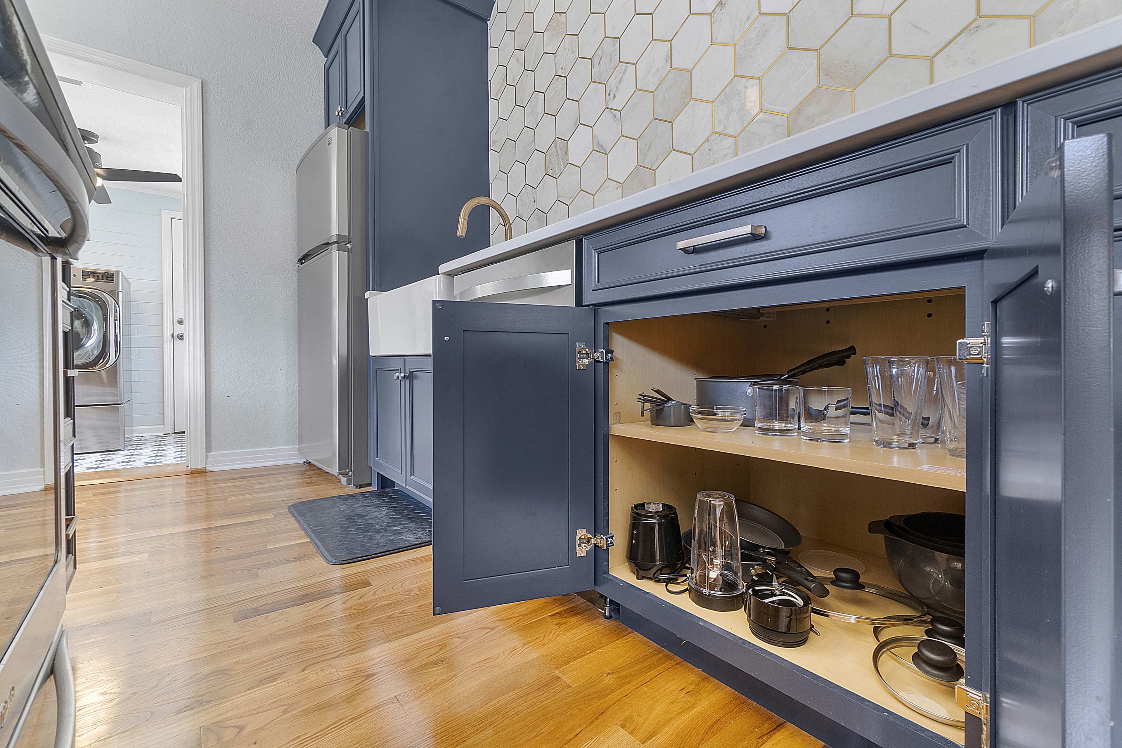 A kitchen with navy blue cabinets and a hexagonal tile backsplash, featuring an open cabinet displaying various kitchen utensils and glassware.