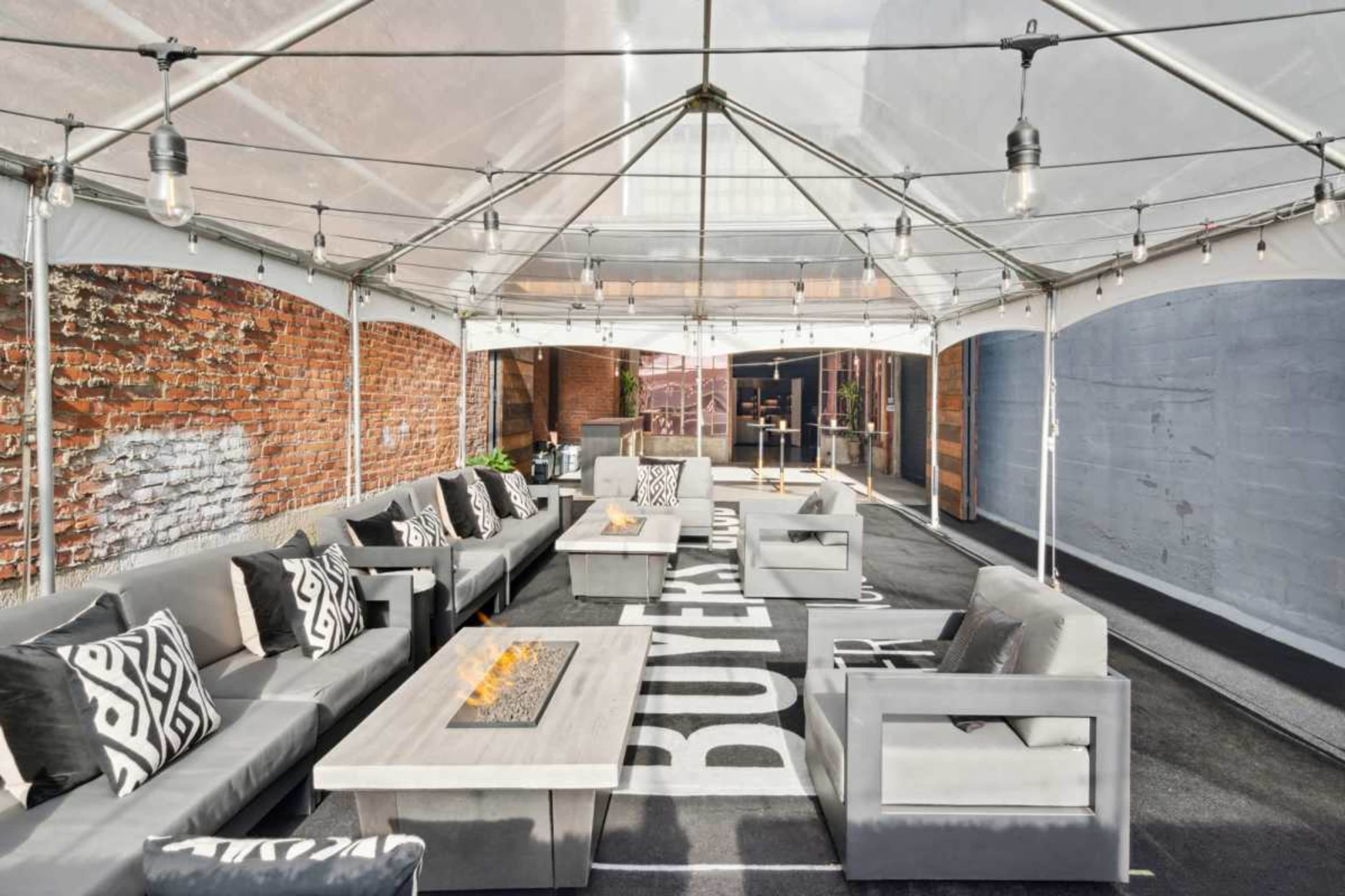 The image shows a spacious tented area furnished with gray sofas and tables, featuring a flame centerpiece, against a backdrop of brick walls.