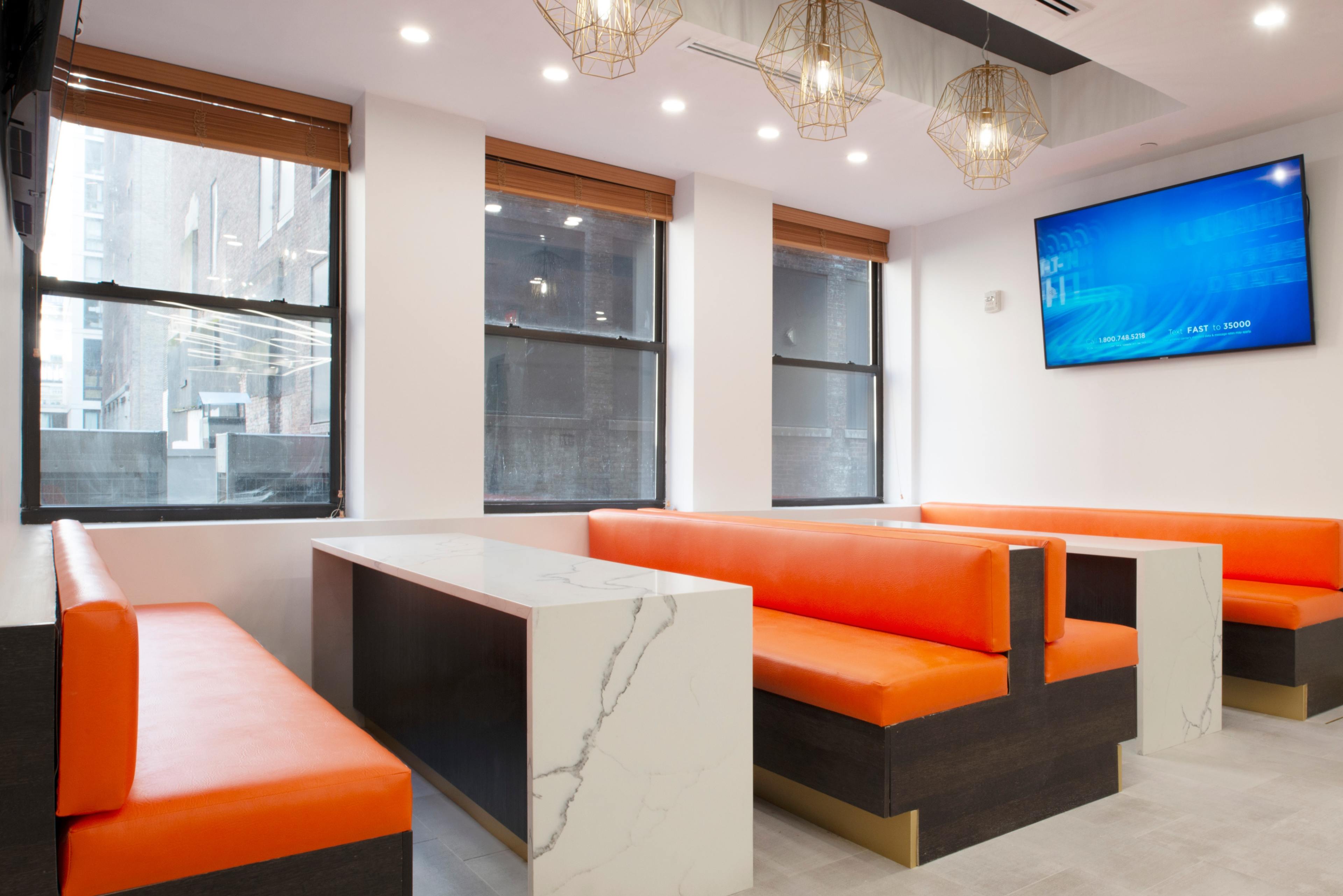 A modern, minimalist dining area featuring orange upholstered benches, a marble-topped table, and large windows with wooden blinds.