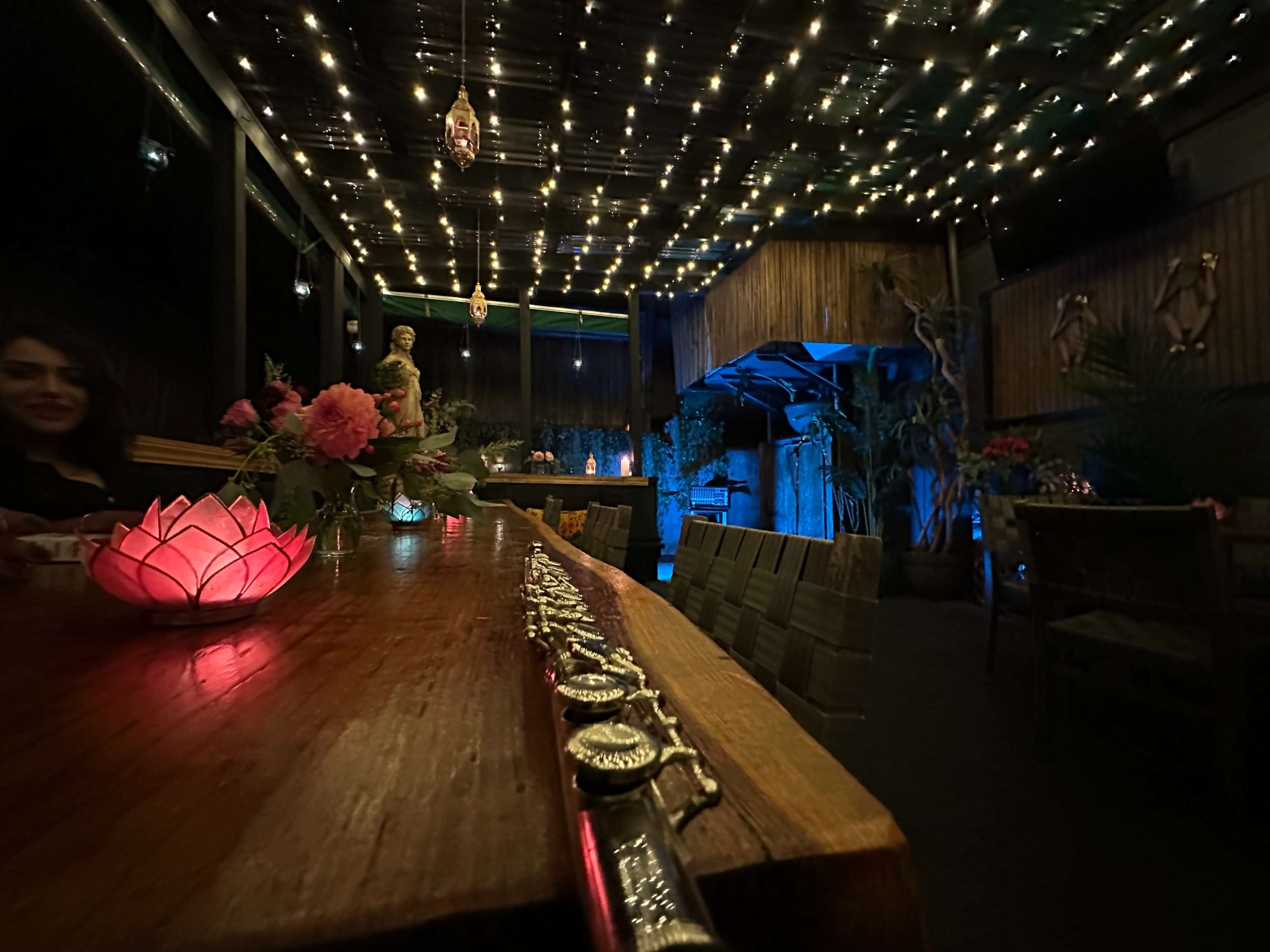 The image shows the interior of a dimly lit restaurant with a long wooden table adorned with flowers and decorative lighting, creating a cozy atmosphere.