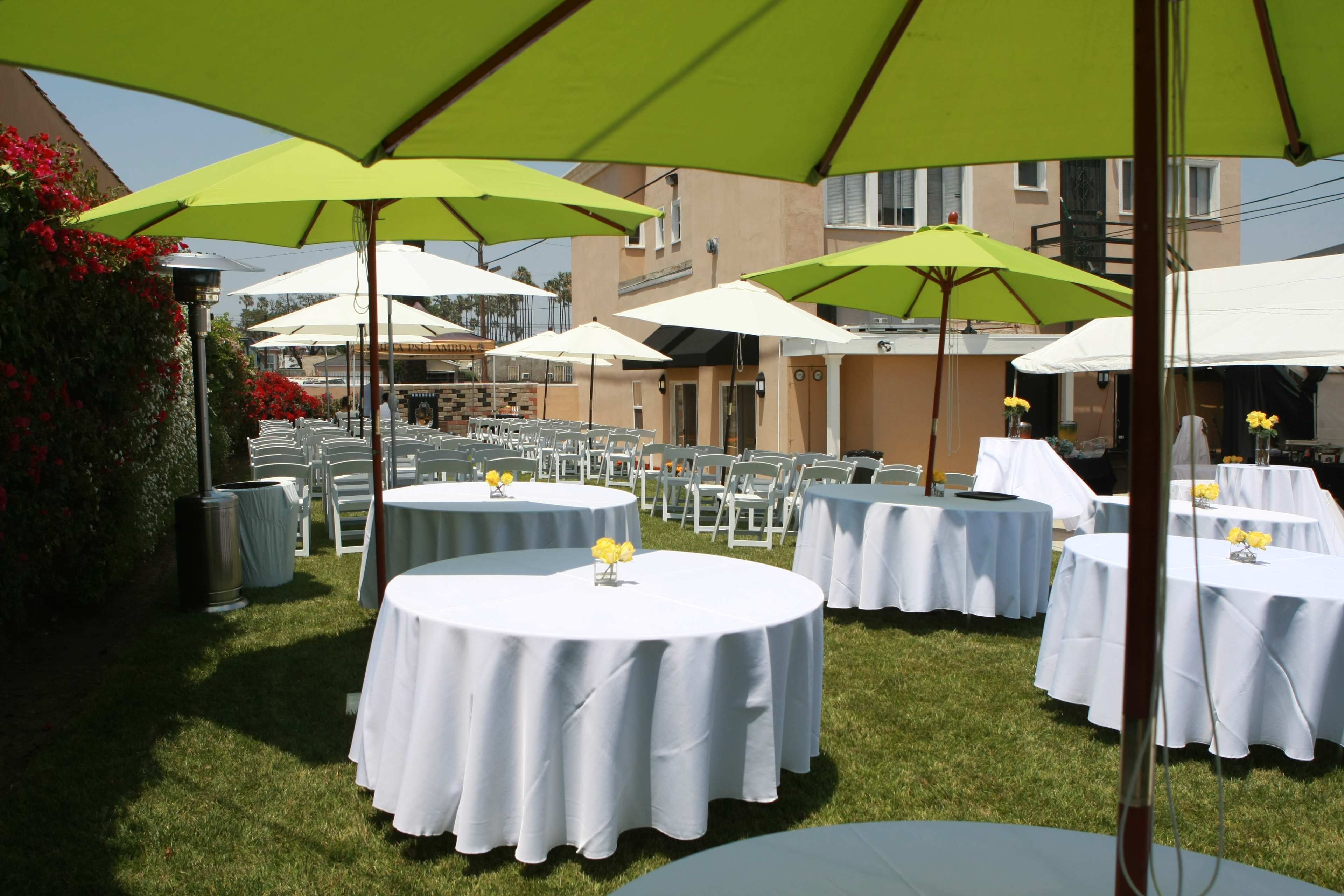 A setup of outdoor tables with white tablecloths and green umbrellas in a grassy area, surrounded by buildings and potted plants.