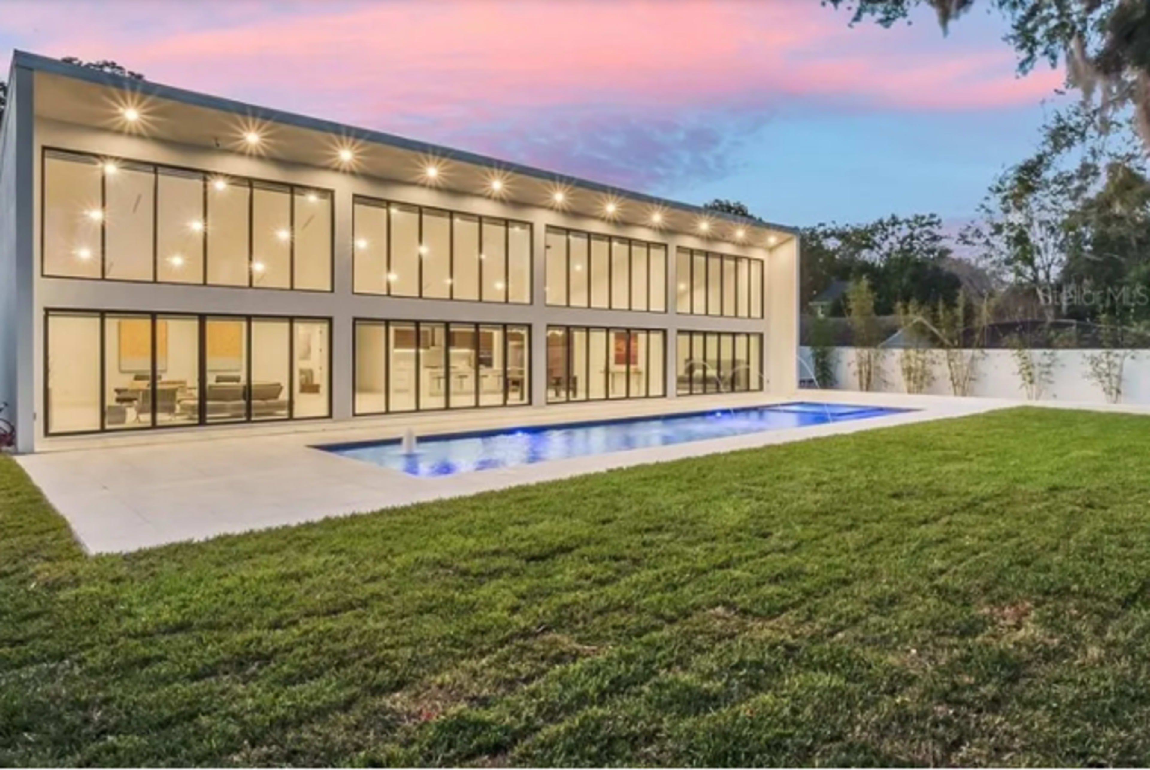 The image shows a modern two-story house with large glass windows and a swimming pool in the backyard, surrounded by green grass.