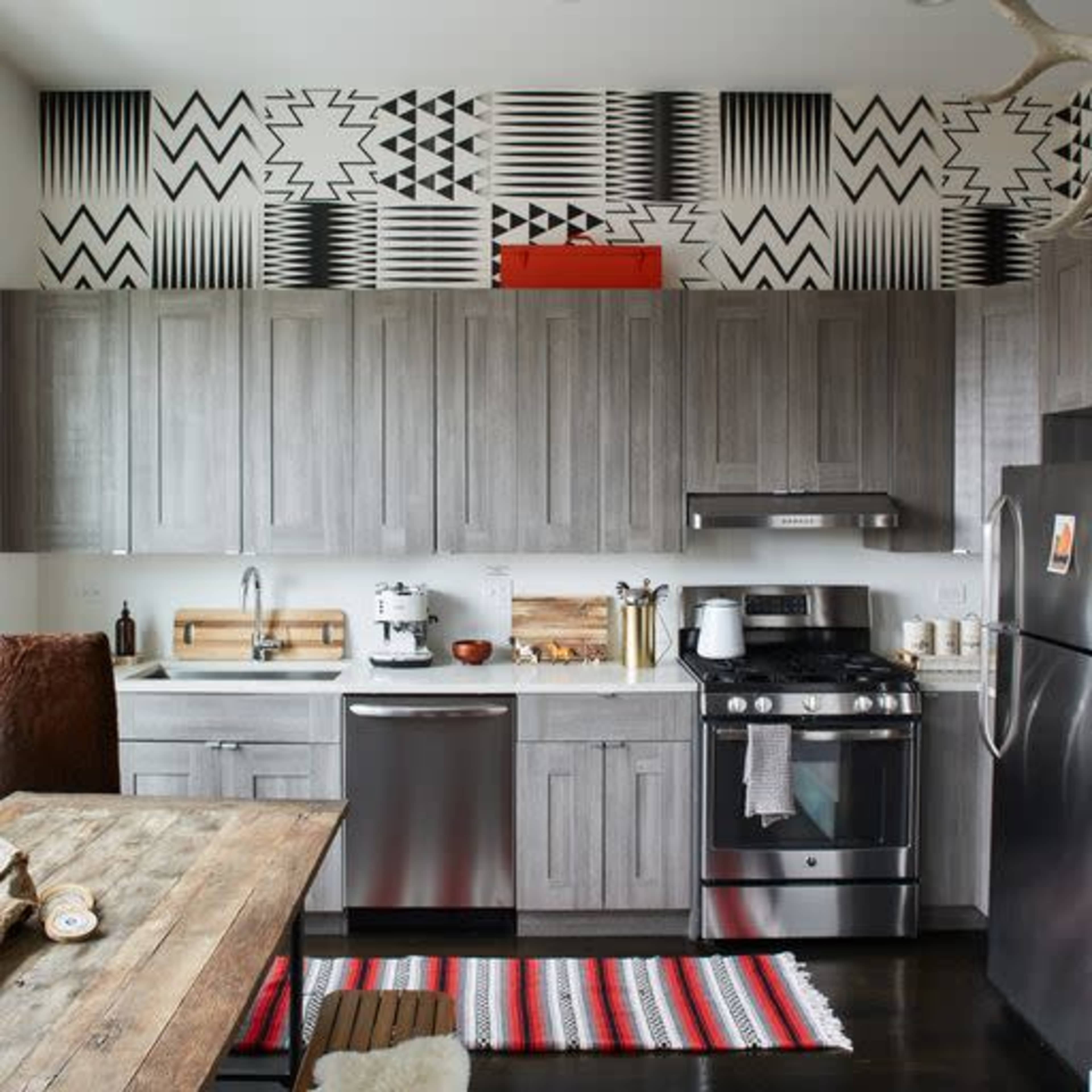 A modern kitchen with gray cabinetry, stainless steel appliances, and a patterned backsplash, accompanied by a wooden dining table and a colorful rug.