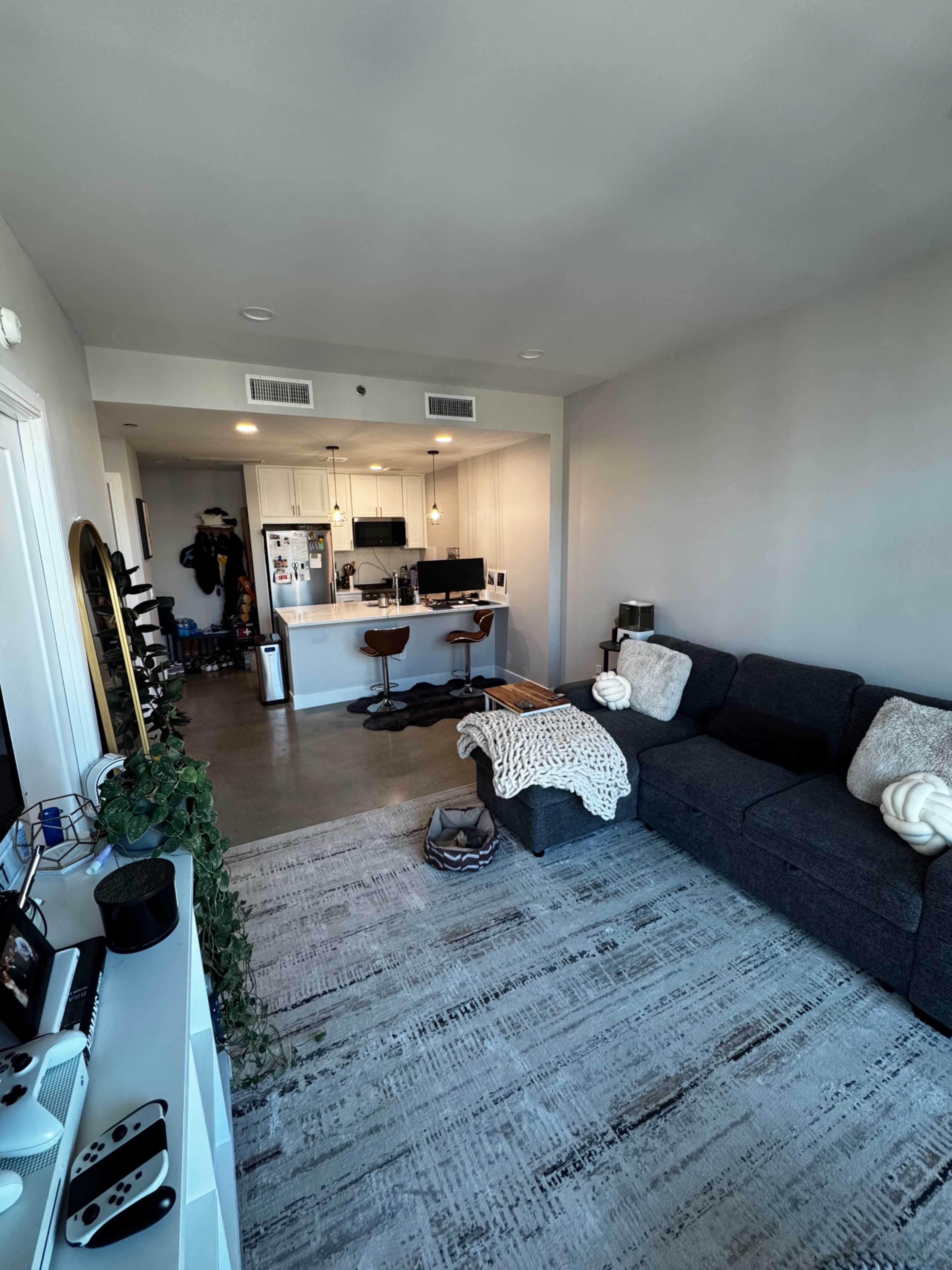A modern living room features a grey sectional sofa, a rug, and a view into an open kitchen with bar stools.