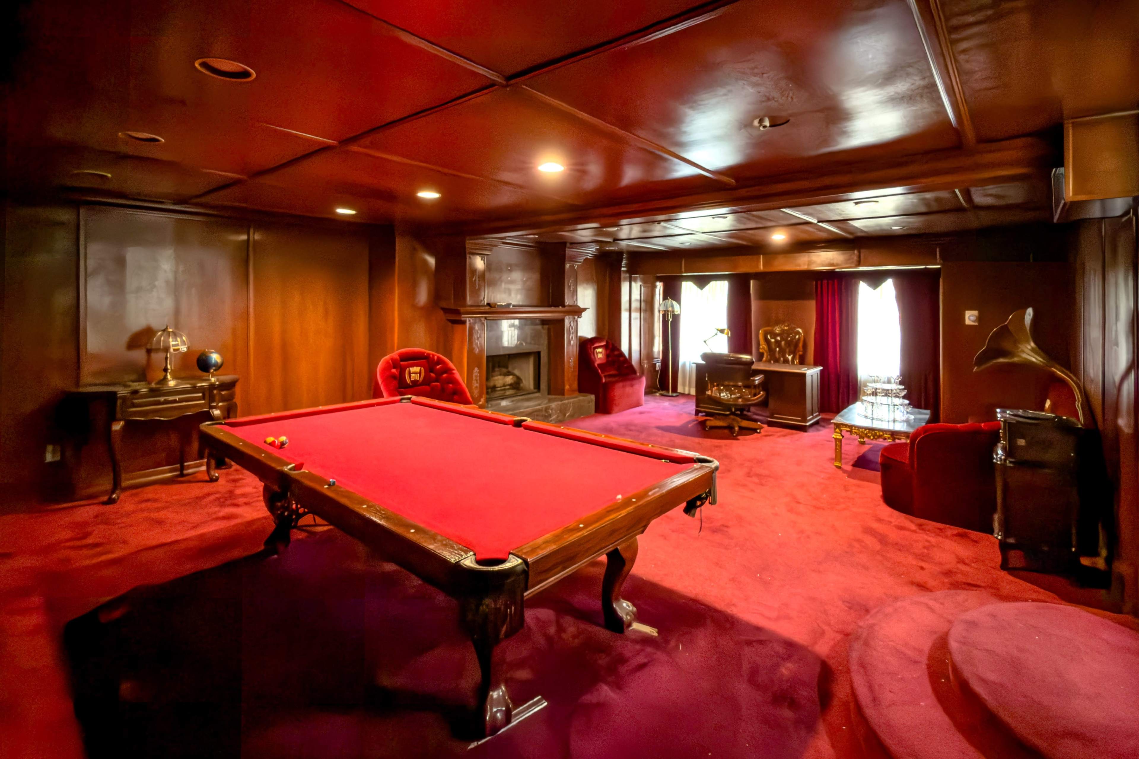 The image shows a dimly lit room featuring a red pool table, plush red carpet, and vintage decor, including a fireplace and an antique gramophone.