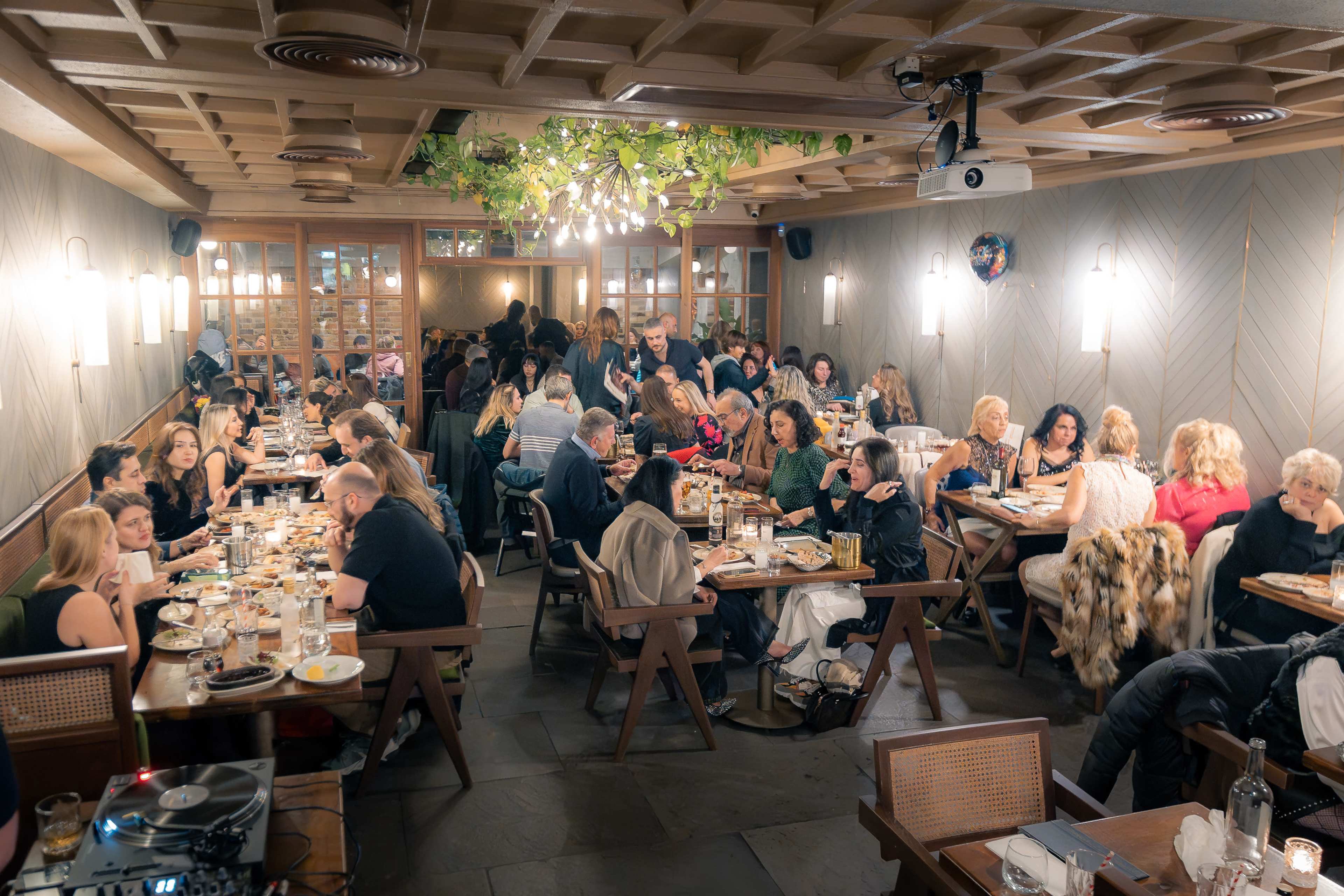 The image shows a crowded restaurant filled with people dining at wooden tables under soft lighting.