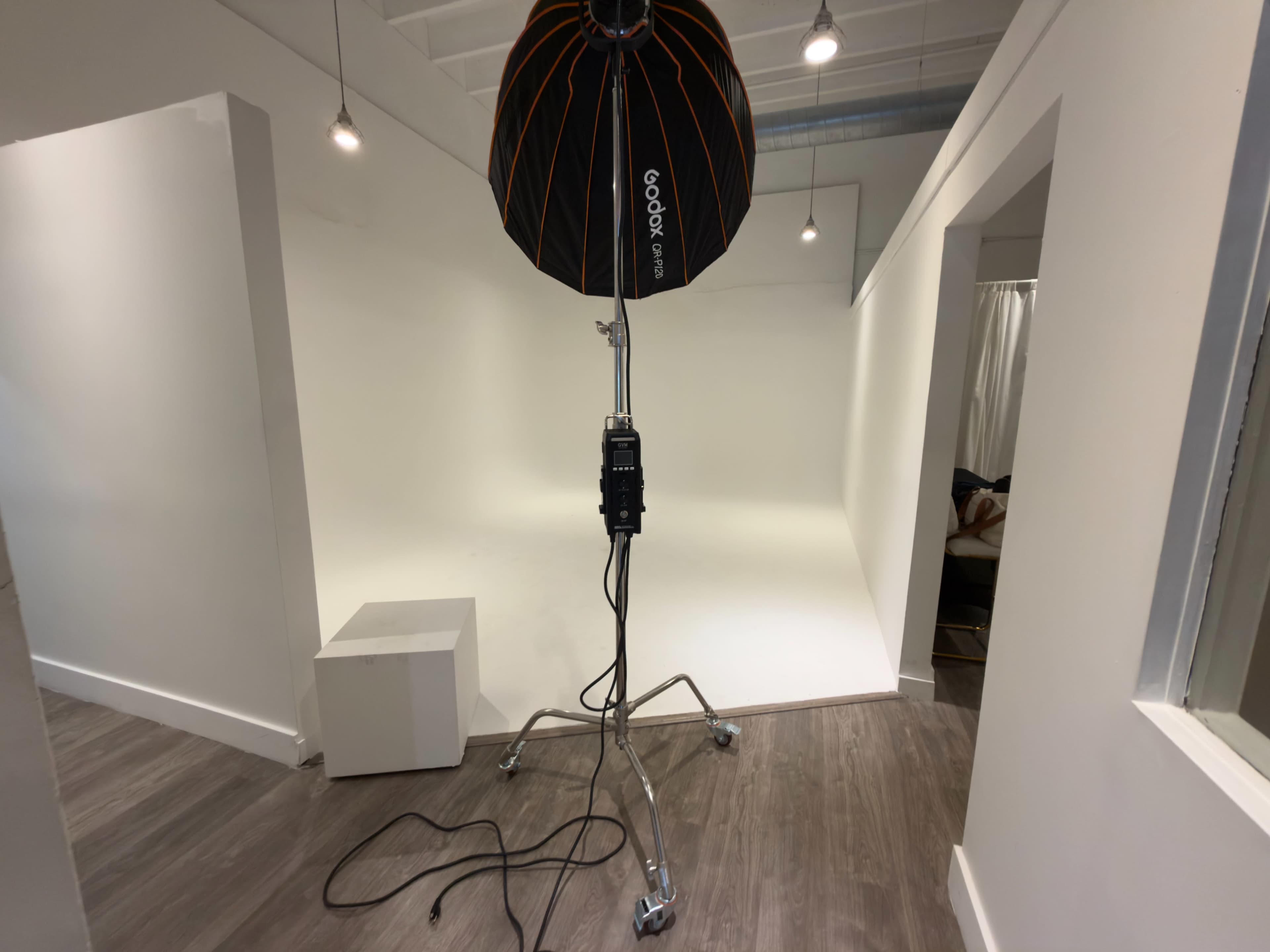 A photography studio features a large light umbrella on a stand facing a plain white backdrop, with a small white pedestal nearby.