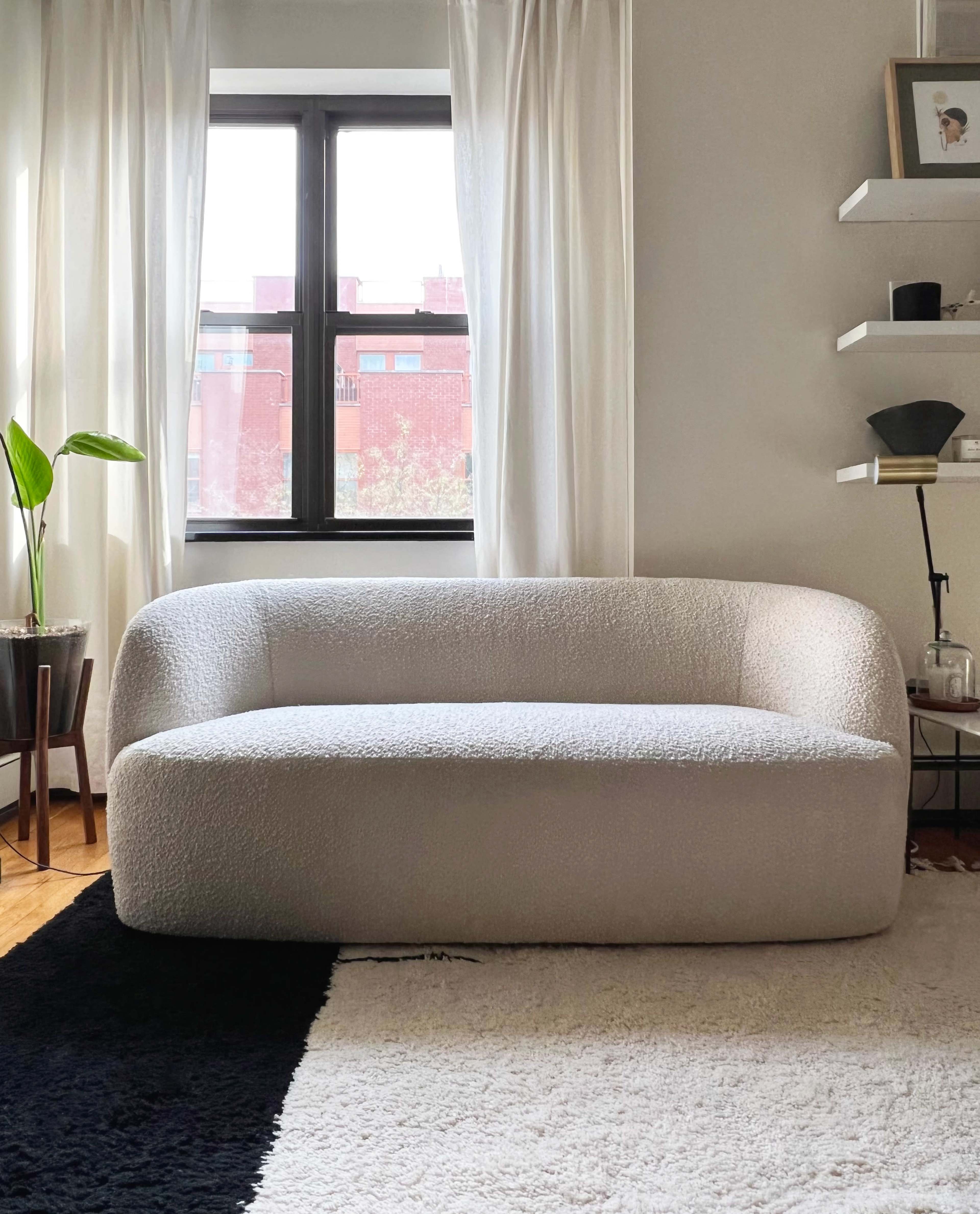 A light-colored, curved sofa is positioned in a room with large windows and white curtains, accompanied by a small plant and a shelf on one wall.
