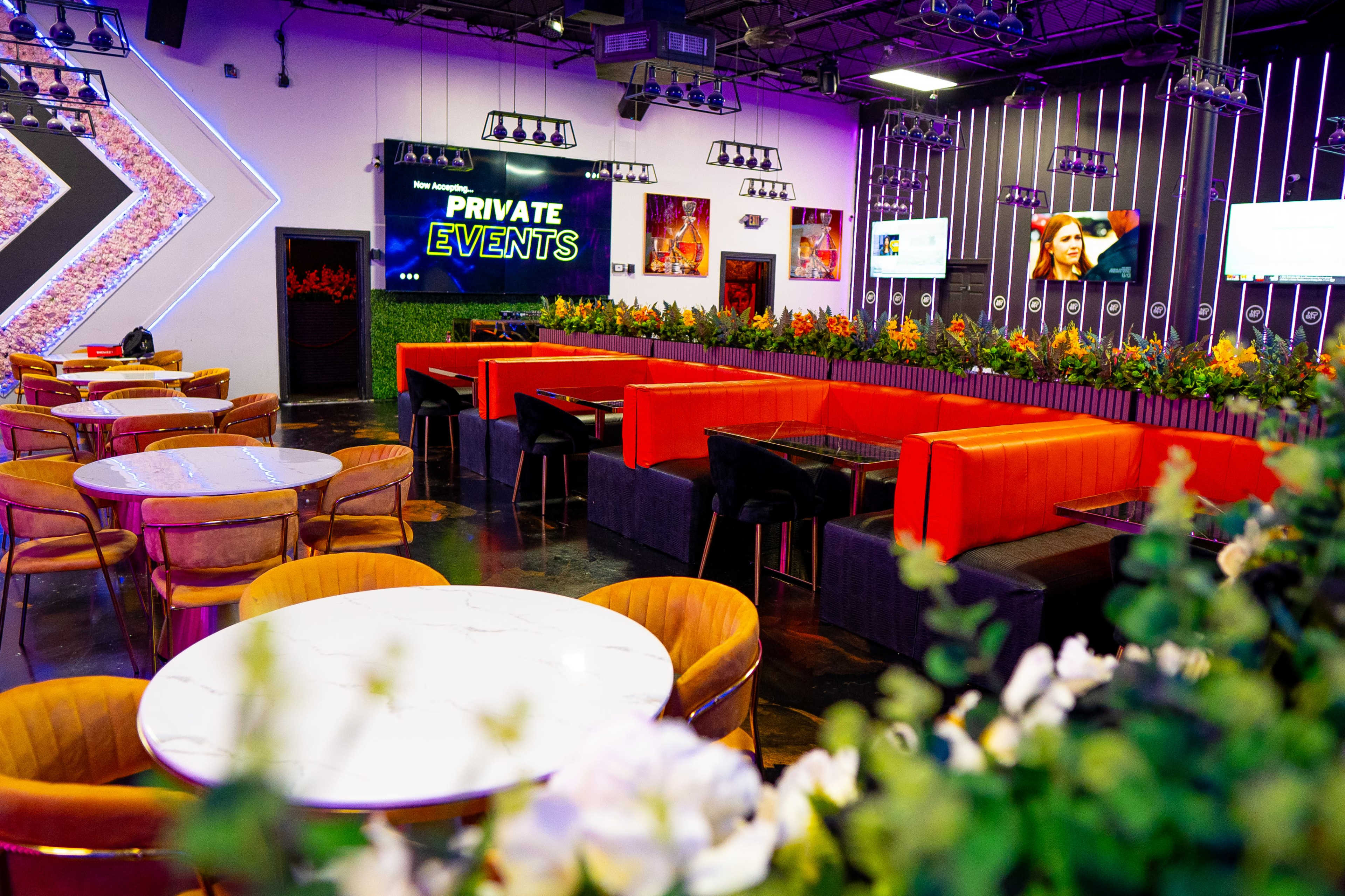 The image shows a modern event space with bright seating areas, circular tables, and colorful floral arrangements under vibrant lighting.