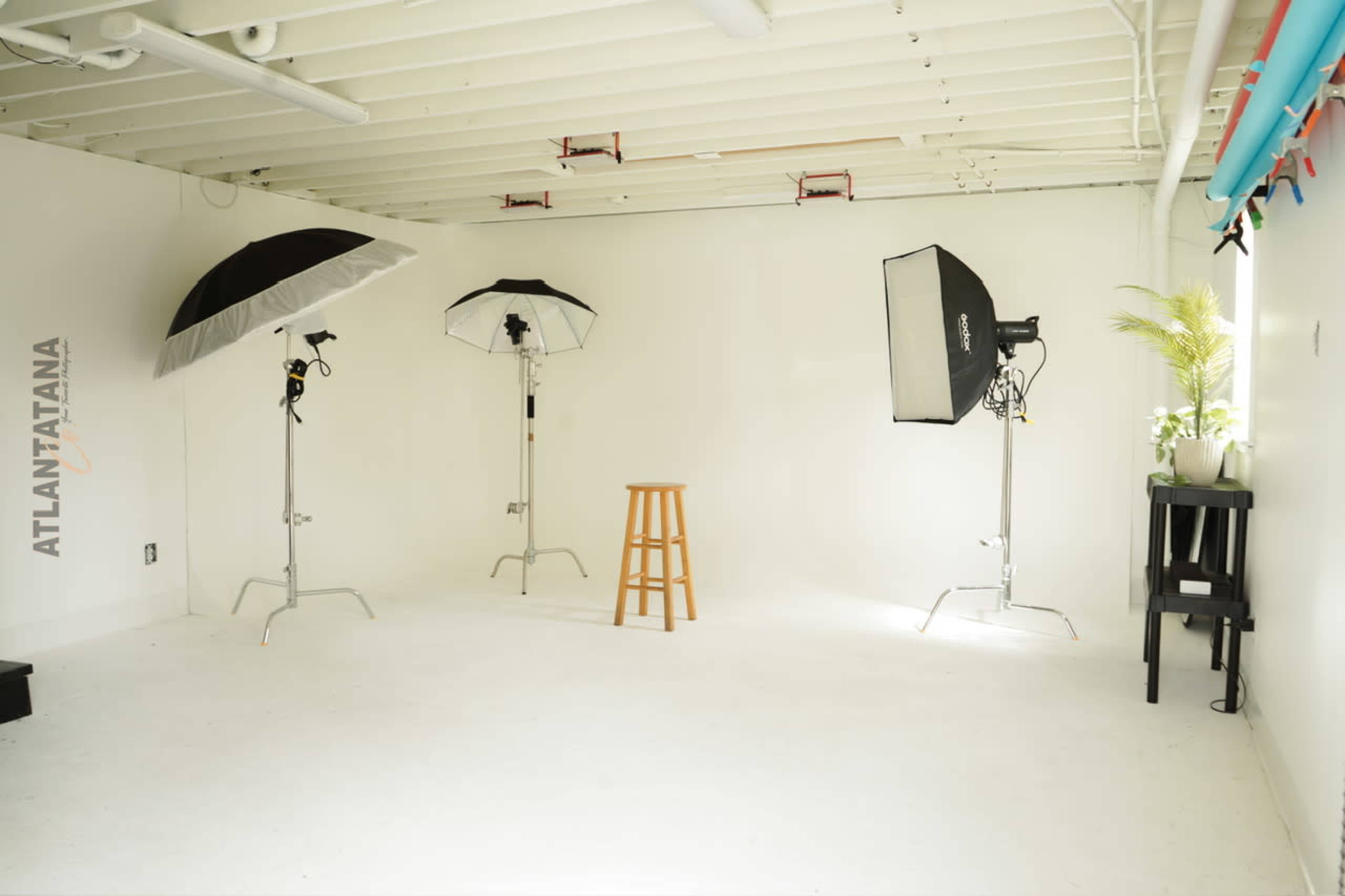 The image shows a well-lit photo studio with three light setups, a wooden stool in the center, and white walls.
