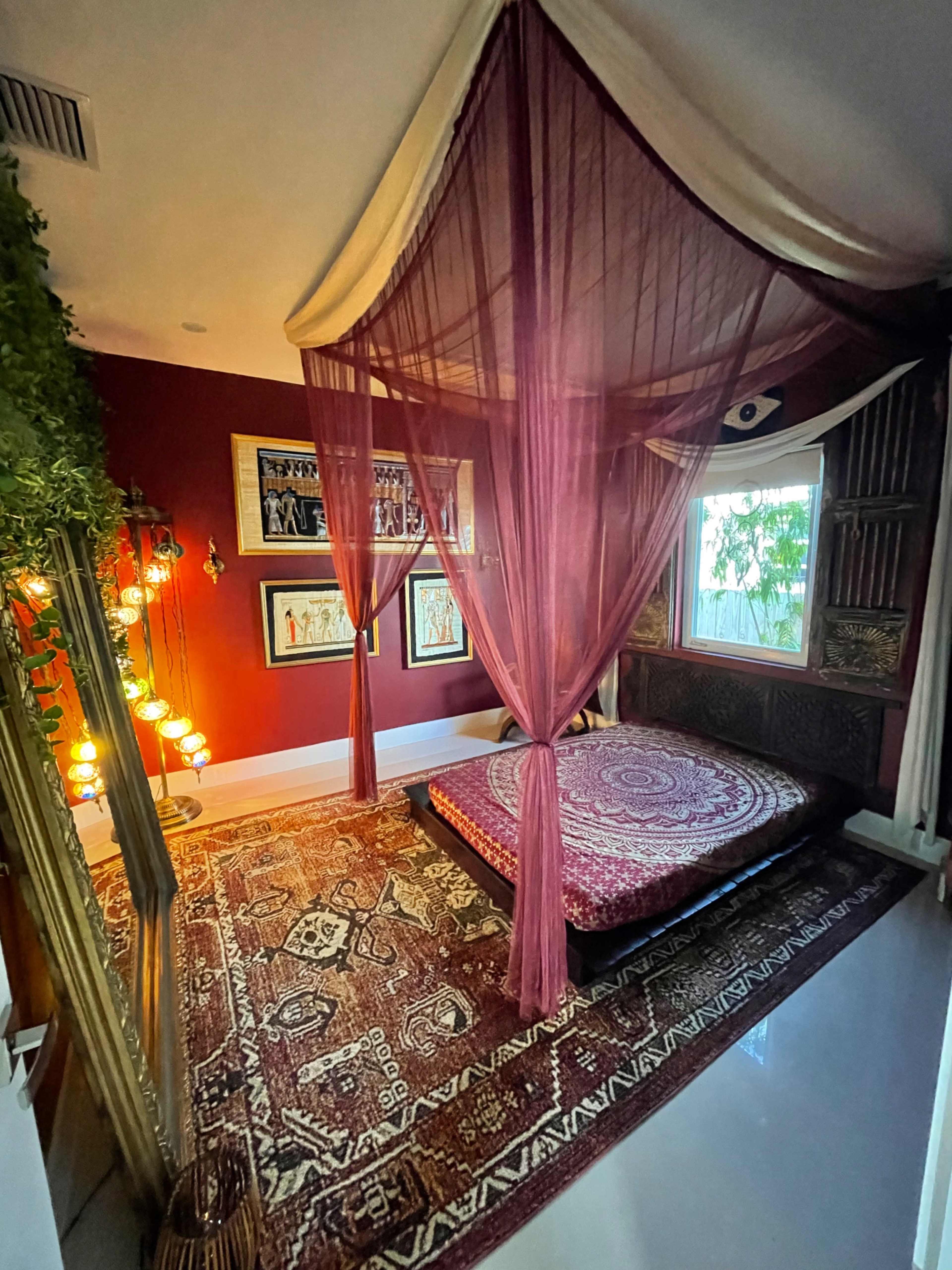 A bedroom features a large, round patterned rug beneath a low, circular mattress draped with sheer curtains, surrounded by decorative wall art and ambient lighting.