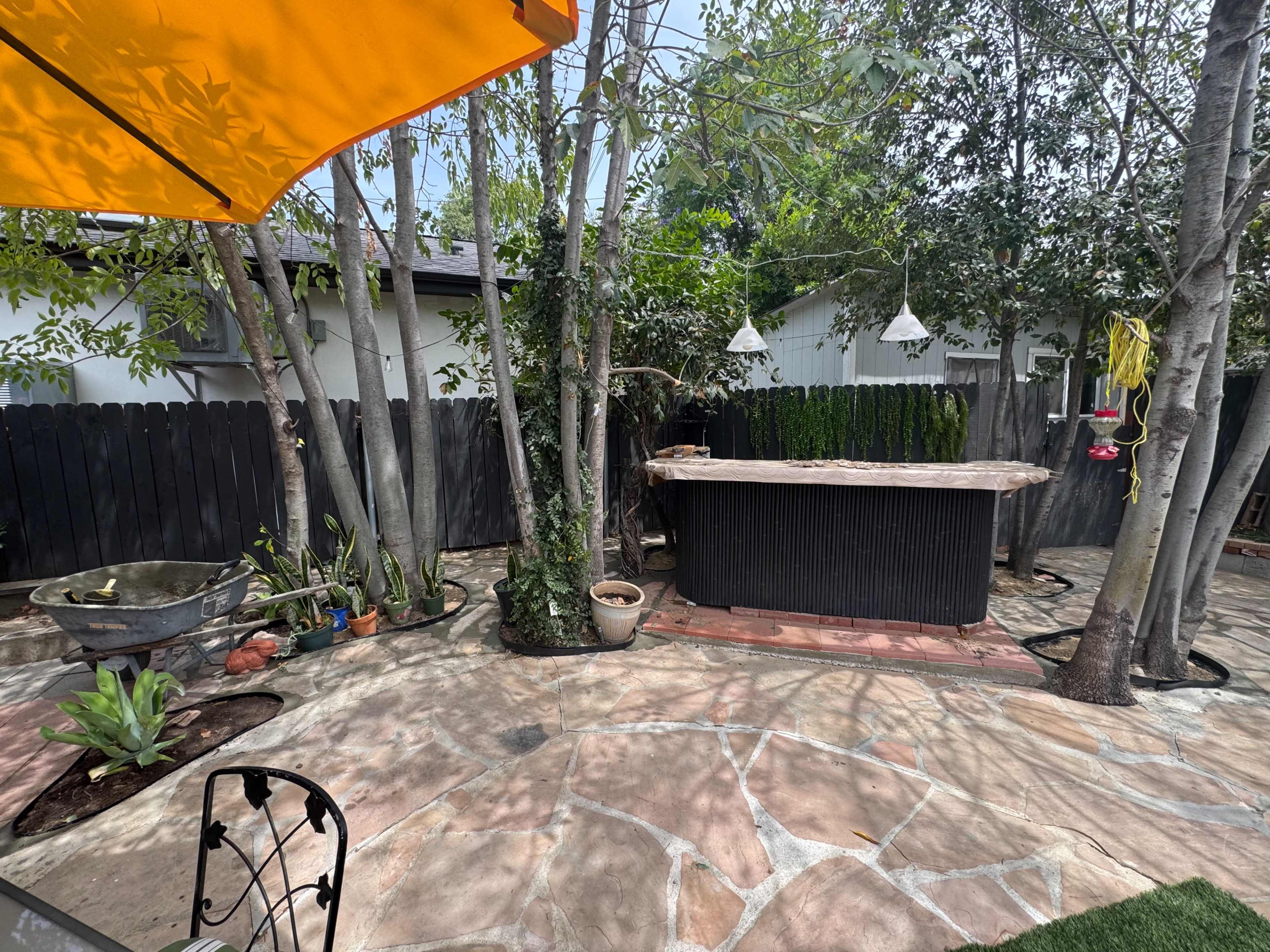 The image shows a backyard with a stone-paved patio, an outdoor bar made from wood, and several trees and plants surrounding the area.