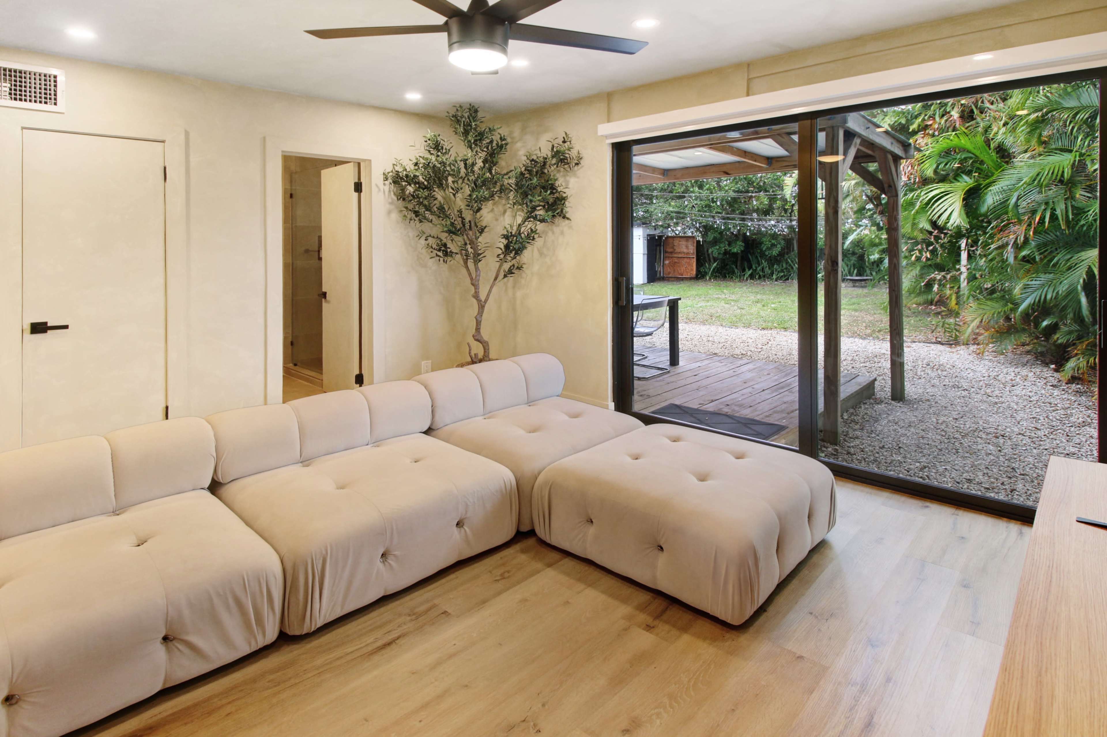 A modern living room features a large beige sectional sofa, a sliding glass door leading to a gravel outdoor area, and a small tree in the corner.