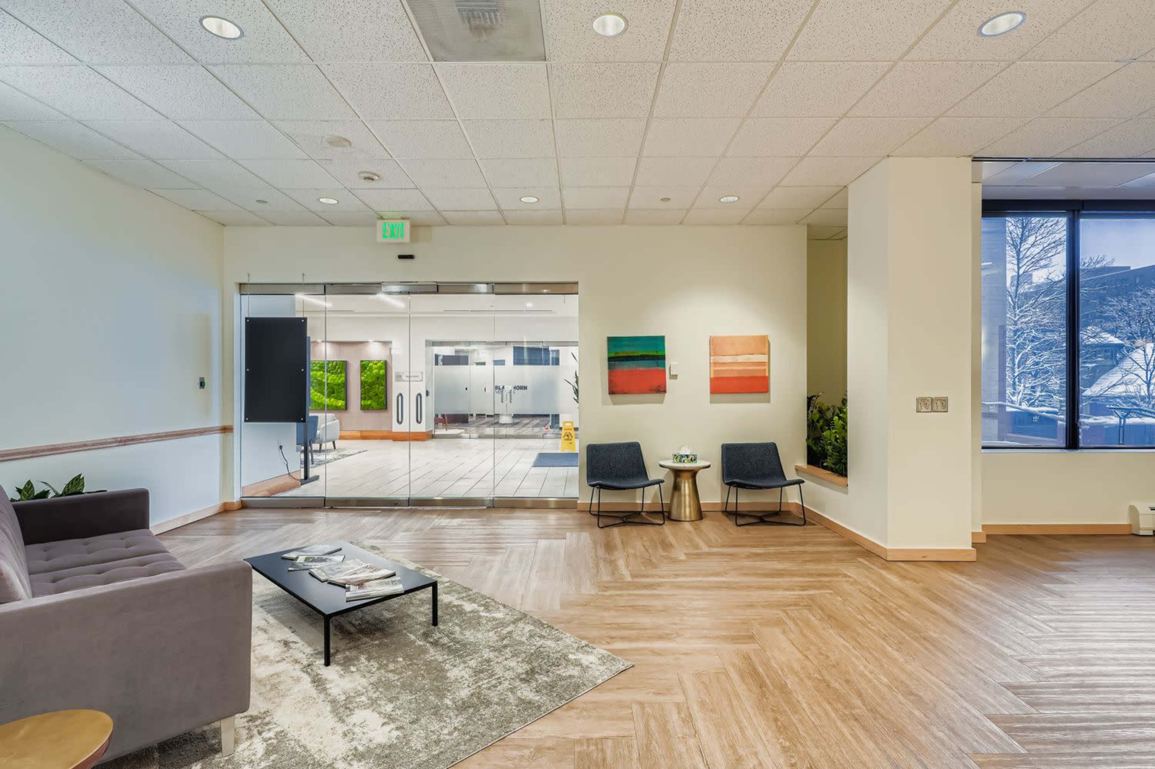 The image depicts a modern indoor lounge area featuring two chairs, a coffee table, and abstract art on the walls, with a glass wall leading to a bright and spacious room.