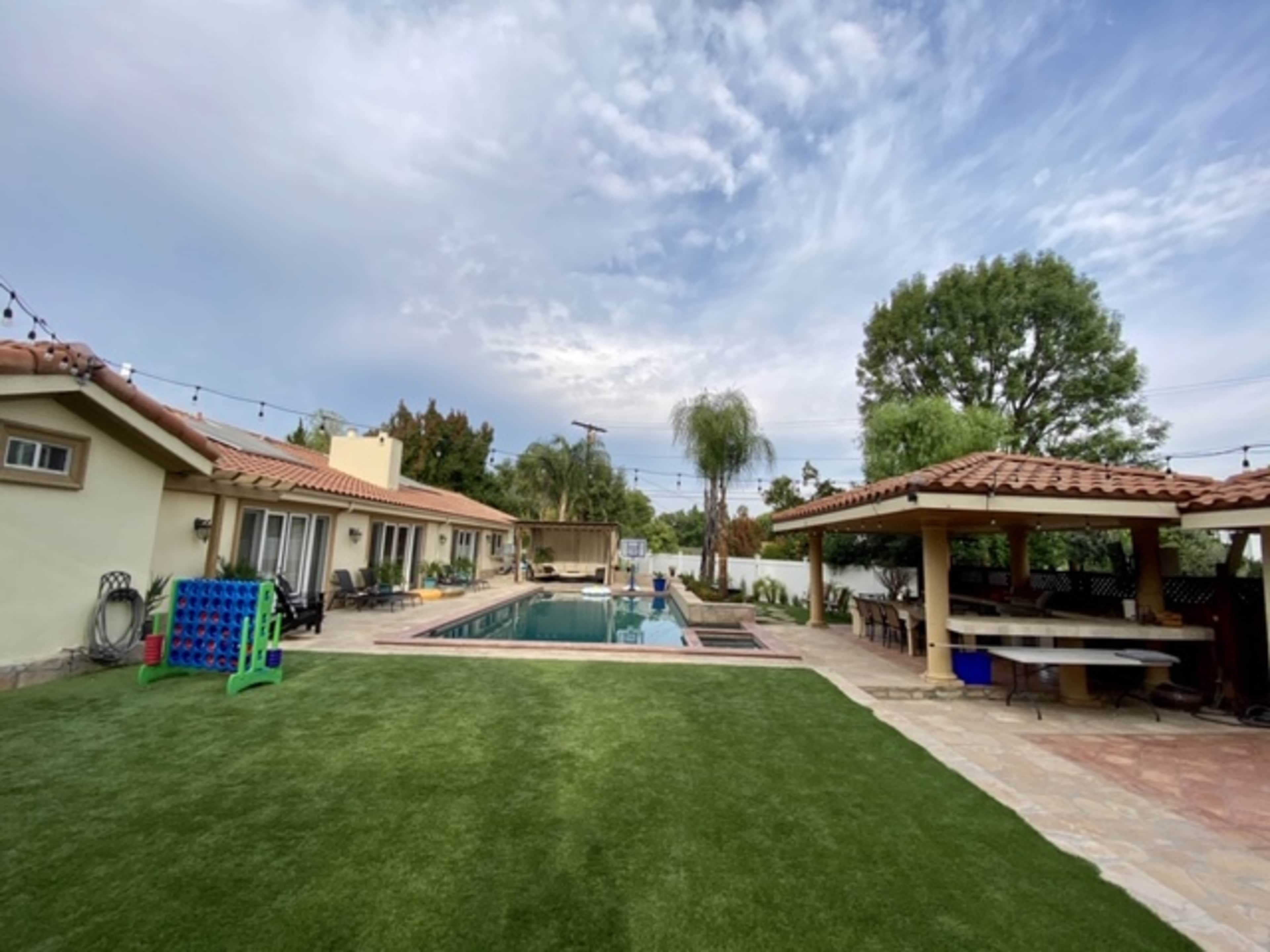 A backyard with a swimming pool, two houses, and a lounge area under a gazebo, surrounded by greenery and patio furniture.