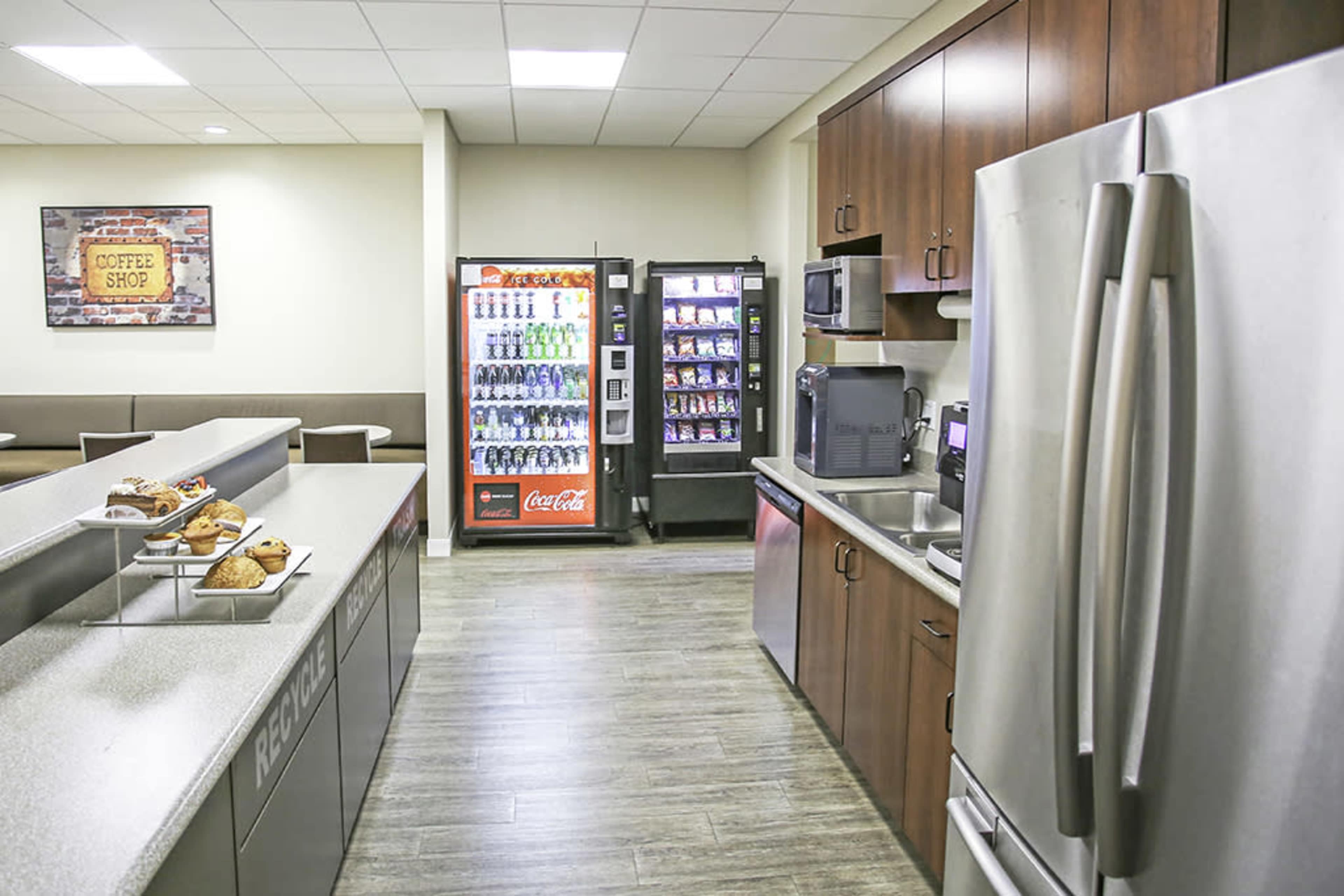 A break room with modern cabinets, a refrigerator, a coffee machine, a vending machine with snacks and drinks, and a counter displaying baked goods.