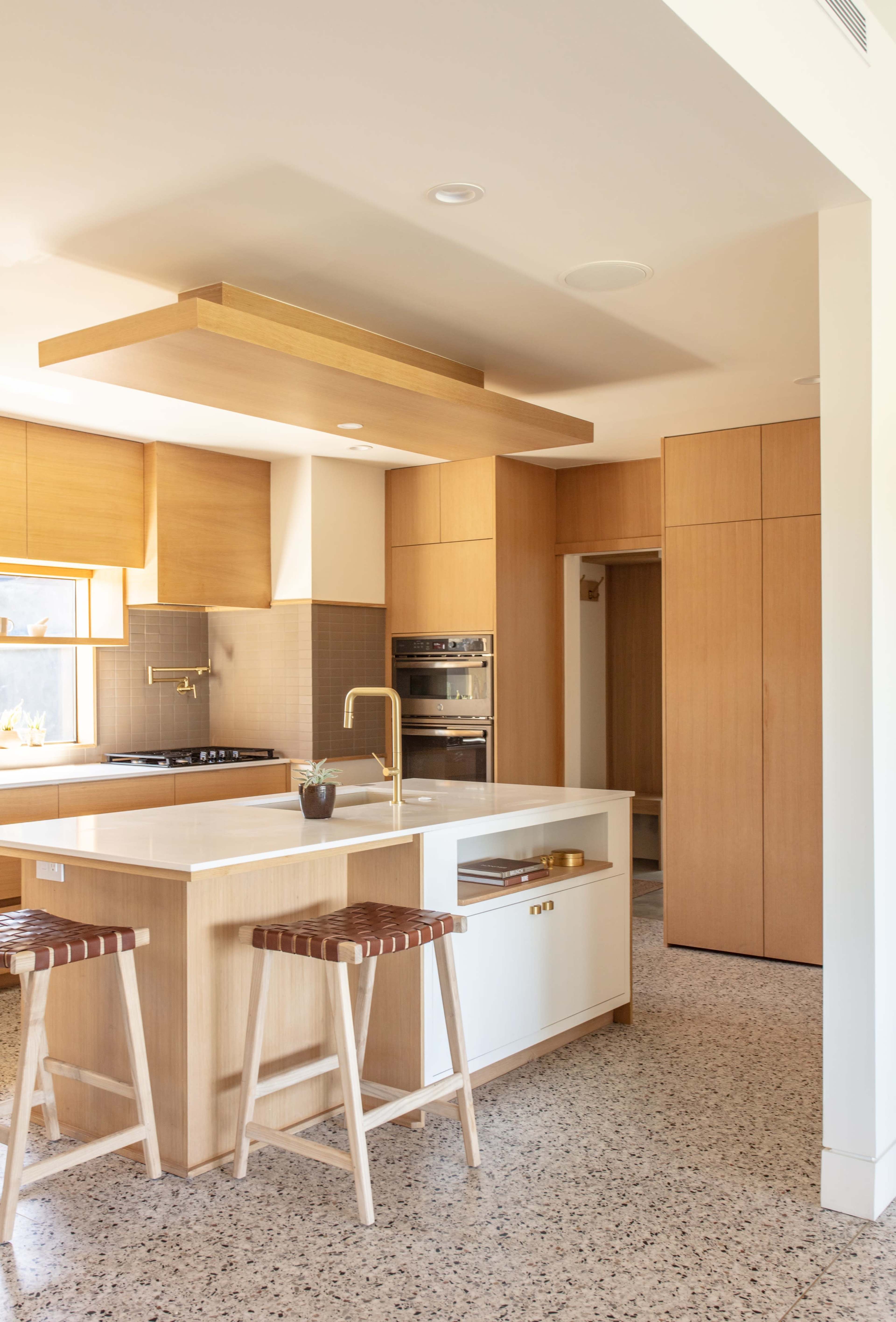 The kitchen features a central island with wooden bar stools, light-colored cabinetry, and modern appliances against a backdrop of speckled terrazzo flooring.