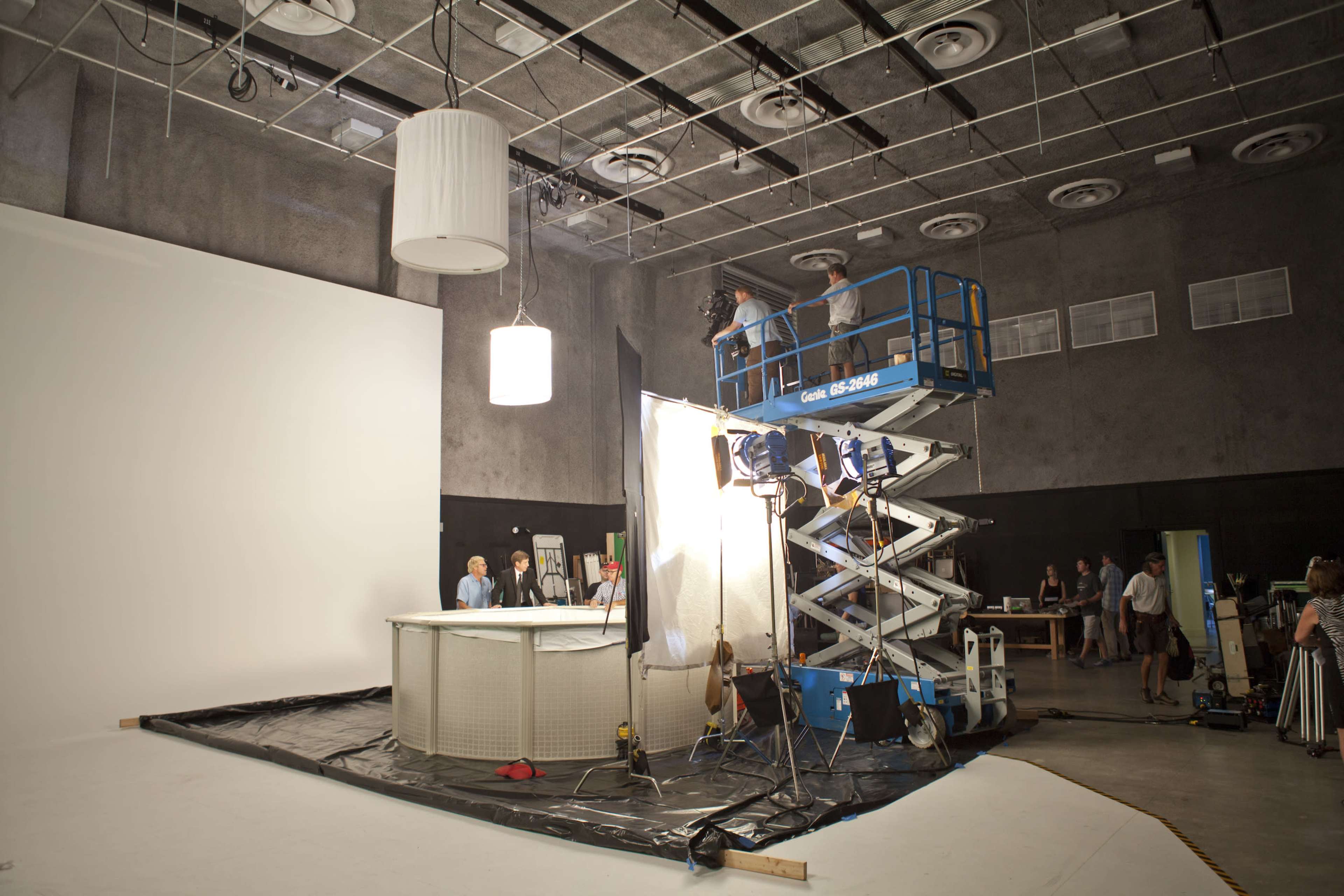 A film crew sets up lighting and equipment in a studio featuring a large white backdrop and a raised platform.